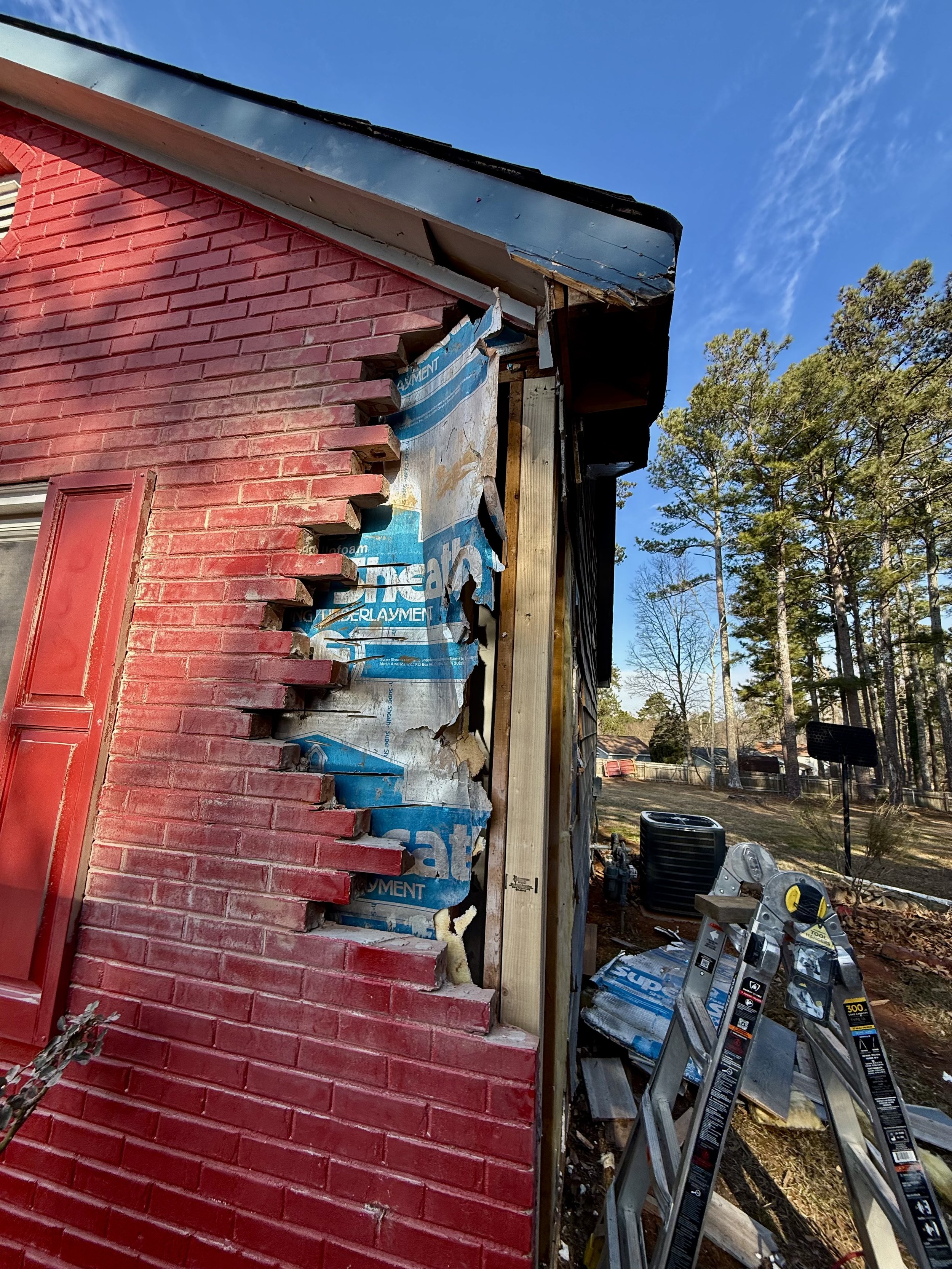 Side of building with damaged red brick exterior, partly torn blue insulation, and construction tools nearby under a clear blue sky.