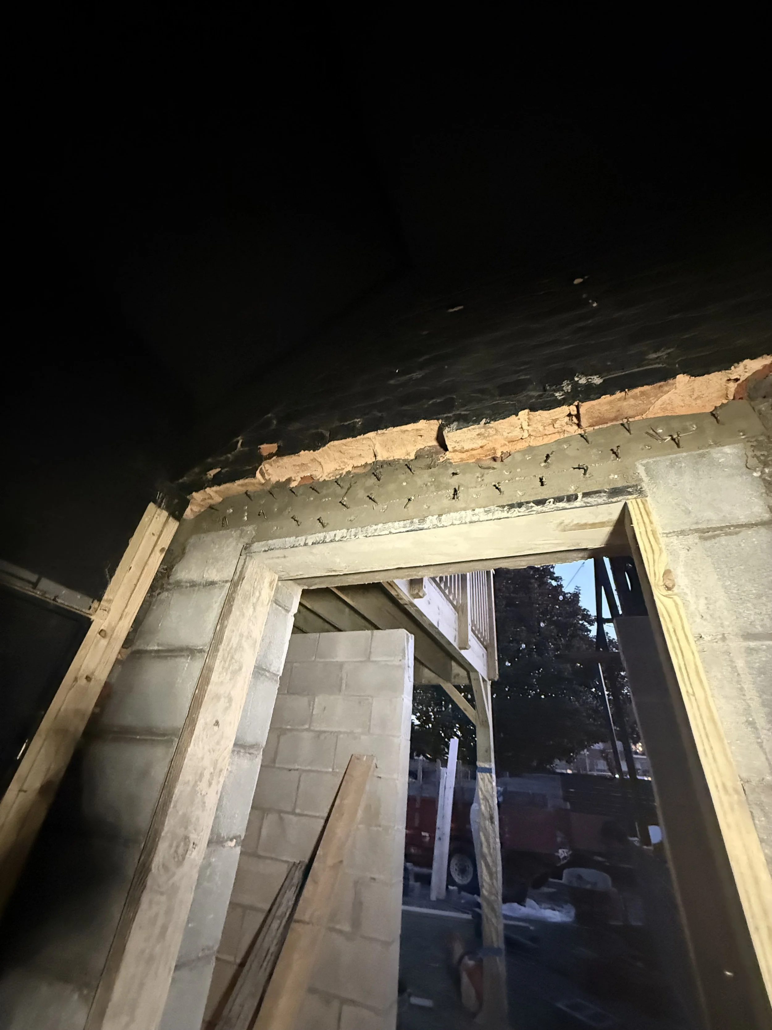 Construction site with brick and wooden framing, showing an open doorway and partial wall, with some outdoor view in the background.