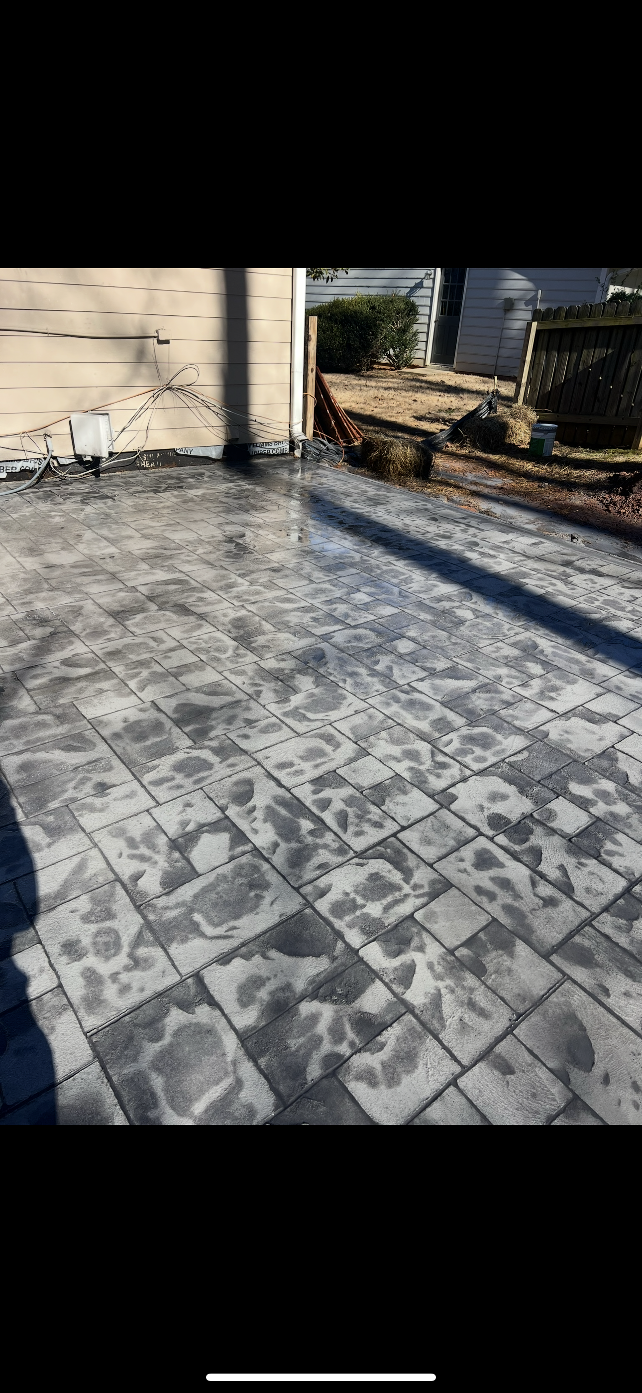 A newly paved concrete patio with a pattern resembling flagstone, wet from a recent cleaning or sealing, in a backyard with houses, bushes, and construction materials in the background.