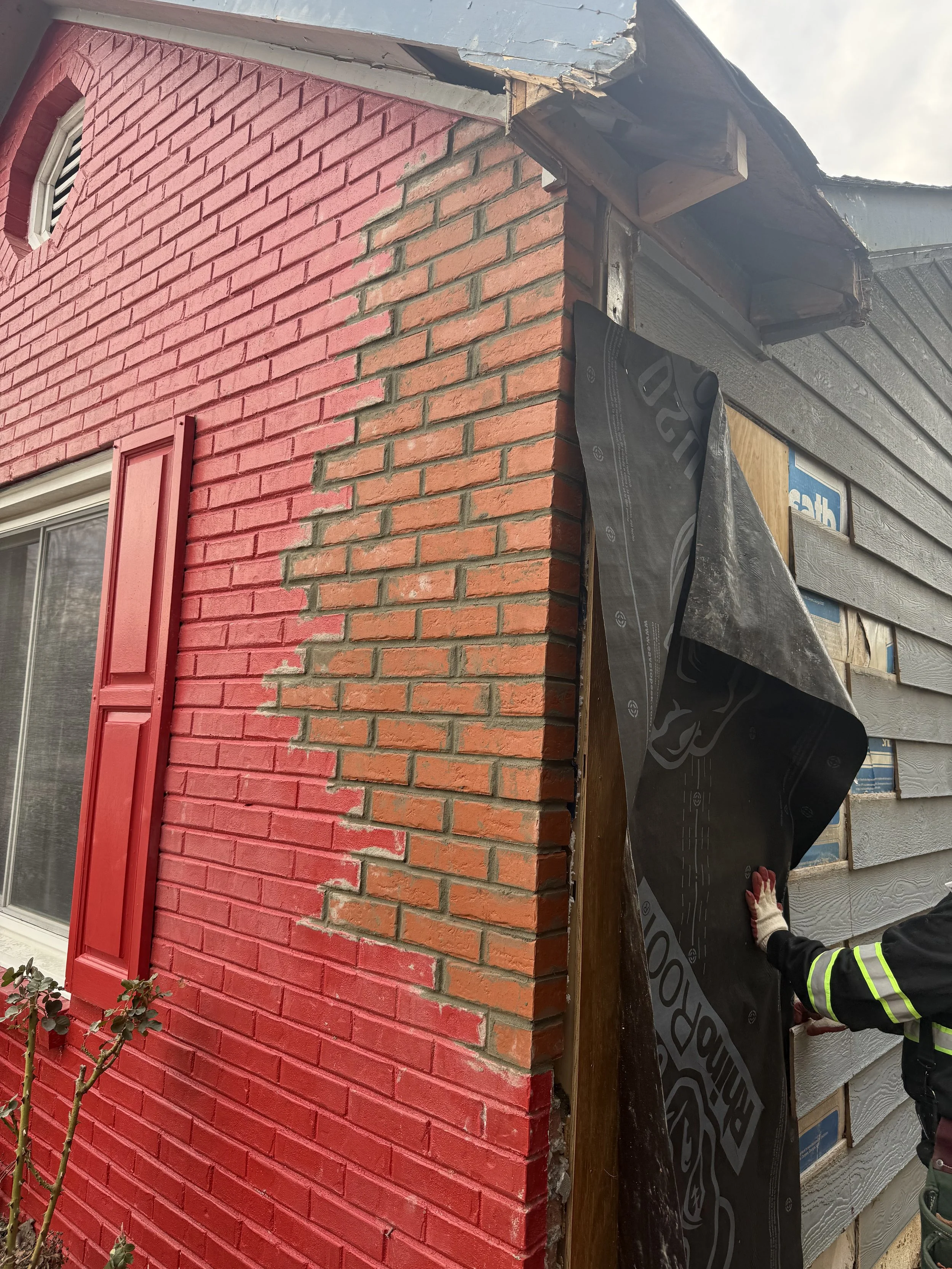 A construction worker installing black house wrap on the corner of a house, which has brick and wood siding, with some of the wood siding covered by the house wrap.