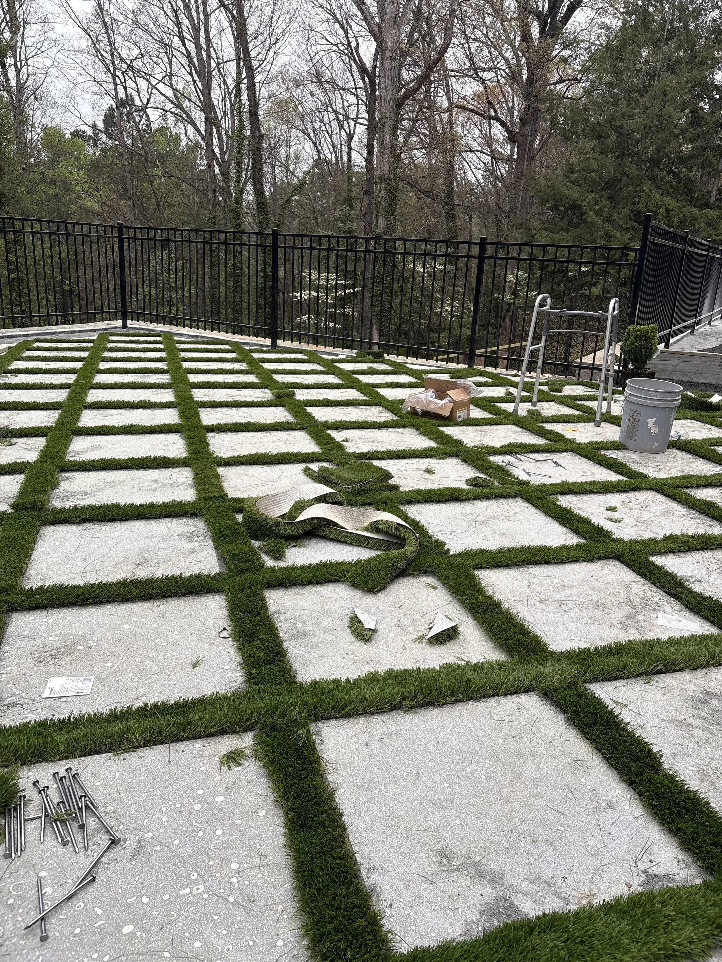 Paved outdoor patio with large concrete tiles, green grass strips in between, and a black metal fence surrounding it. Construction tools and materials are scattered, and some black screws are on the ground.