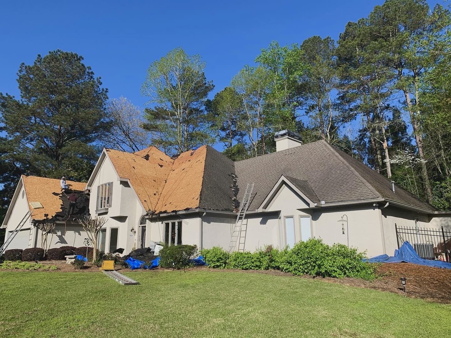 House with roof undergoing repair or replacement, with tenders working on the roof, and surrounded by ladder, gardening tools, and blue tarps on the lawn, with trees and a clear blue sky in the background.