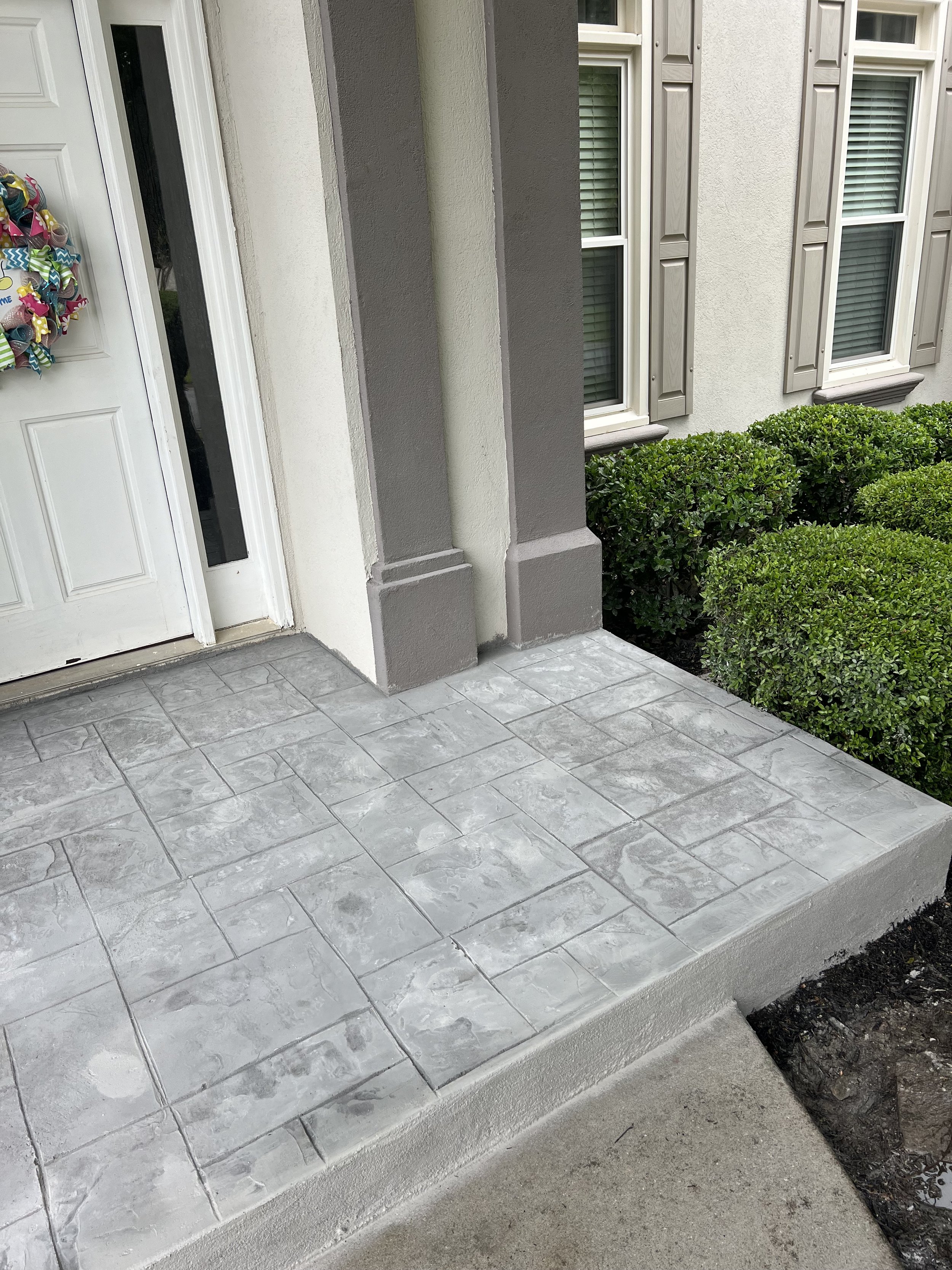 Front porch with grey stamped concrete, bushes, and house windows with shutters.