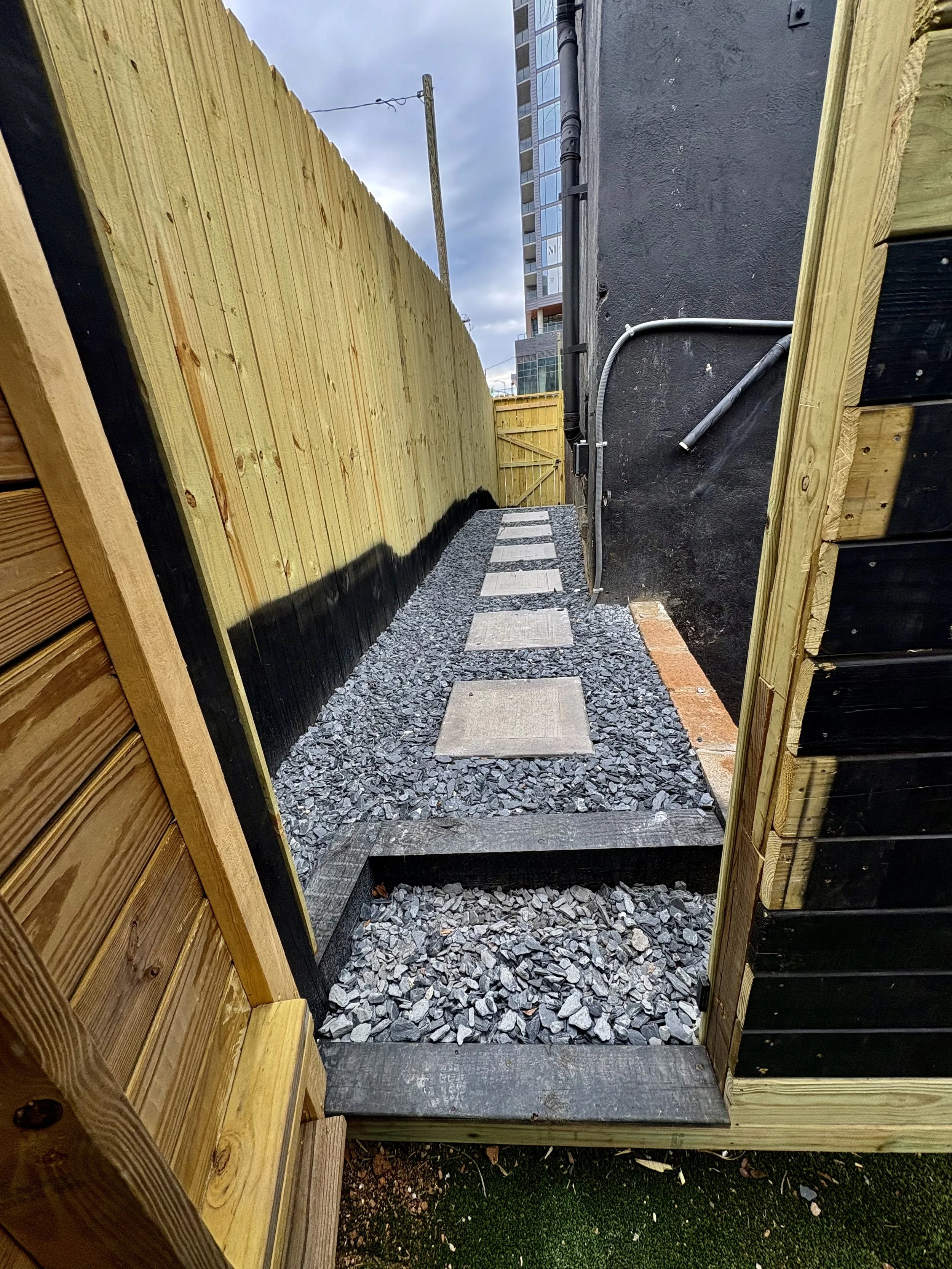 Narrow outdoor pathway with stepping stones, gravel, and wooden borders, flanked by a tall wooden fence on the left and a black wall with exposed pipes on the right, with a tall building and cloudy sky in the background.