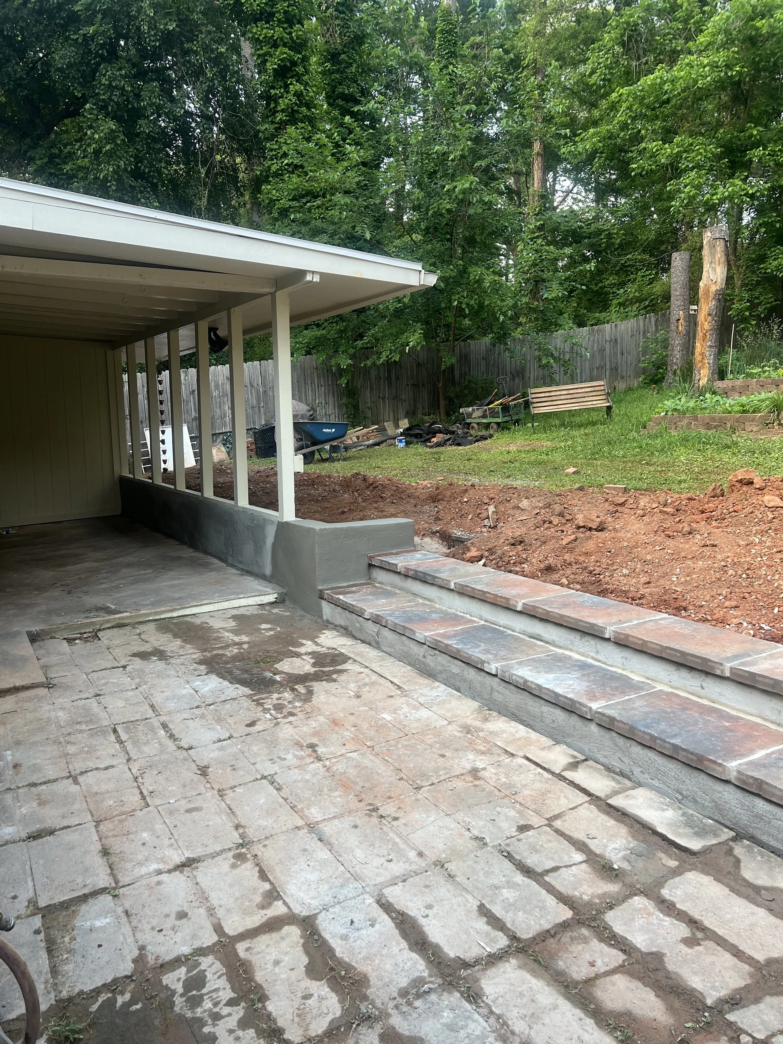Laundry room with brick steps leading outside to a grassy backyard enclosed by a wooden fence, with trees and gardening tools.