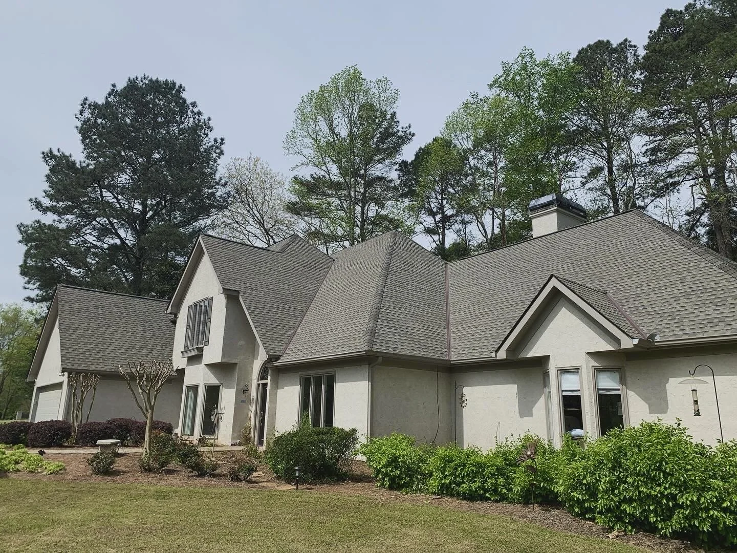 Single-family house with gray roof, white exterior, and a landscaped front yard with shrubs and trees, surrounded by tall trees in the background.