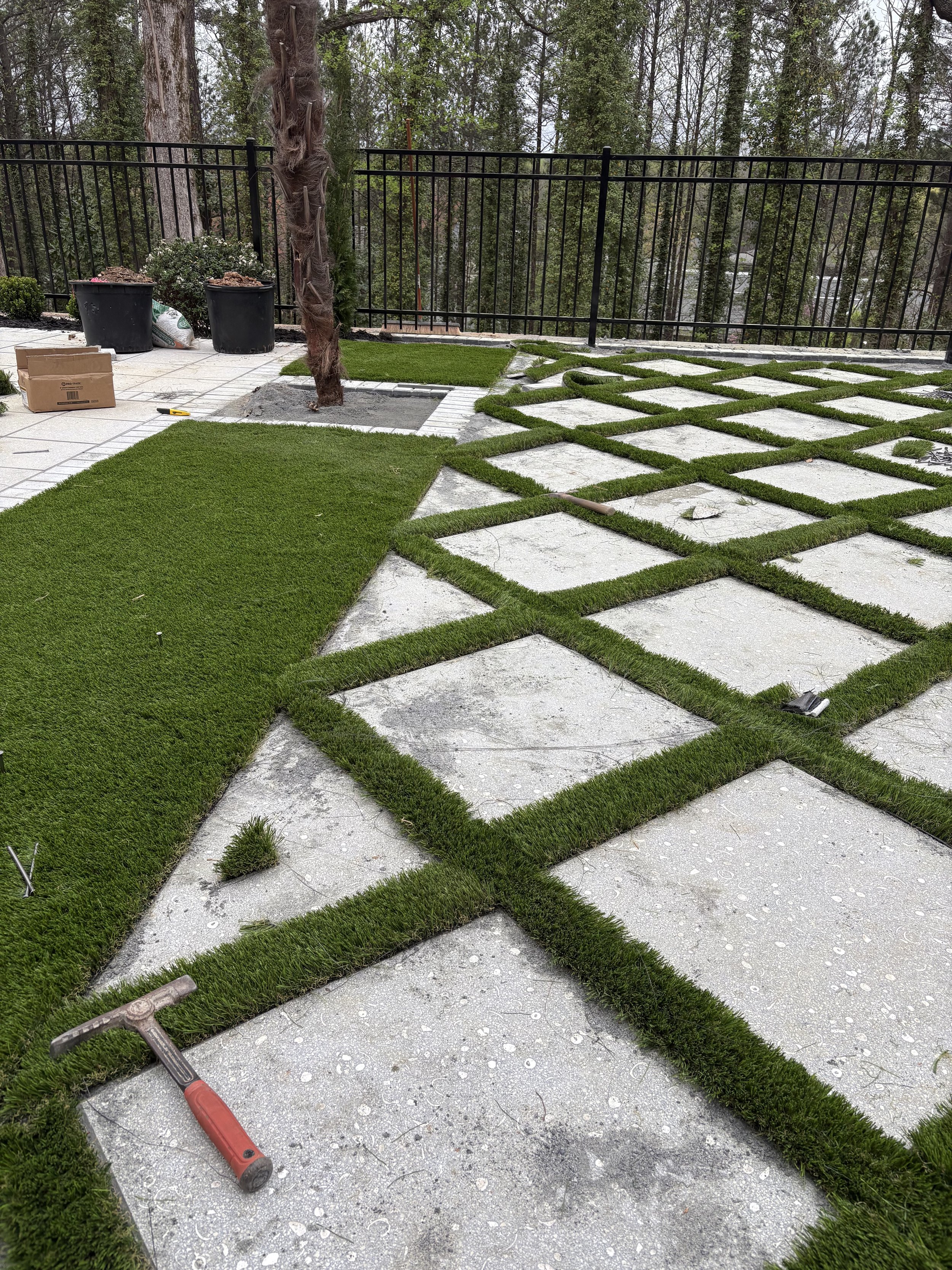 Laying down artificial grass for a decorative outdoor area with a grid pattern on concrete, surrounded by trees and a black metal fence.