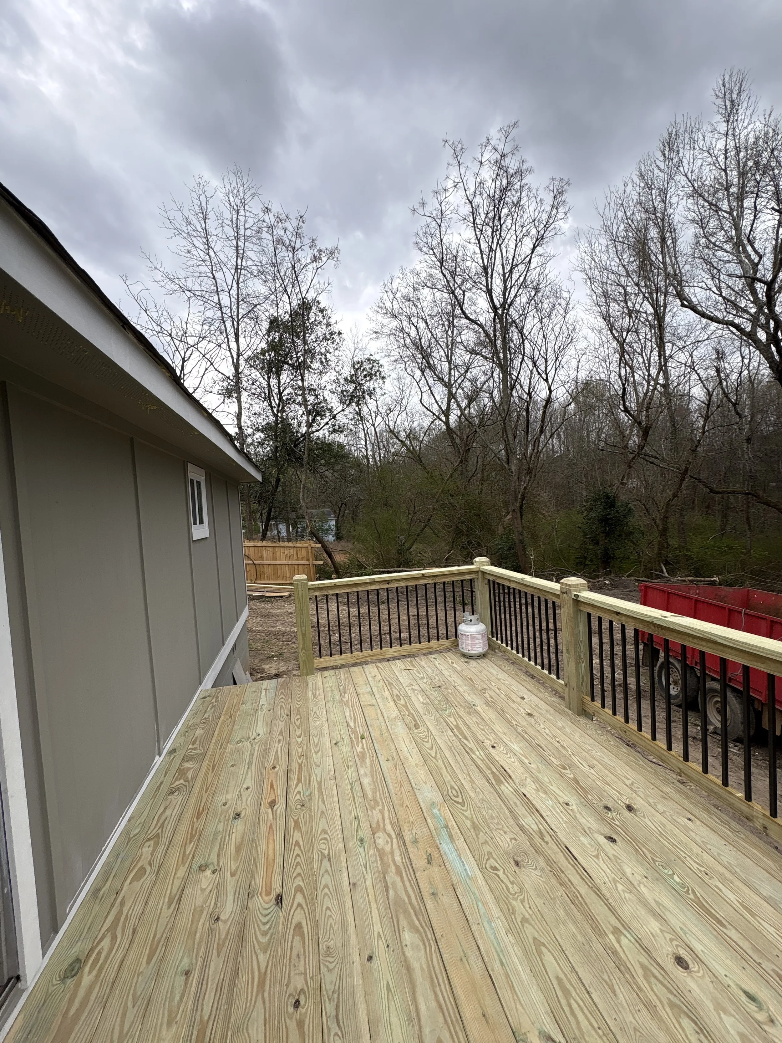 New wooden deck attached to a house with a black railing, a gas tank, and a red truck parked nearby, with leafless trees and a cloudy sky in the background.