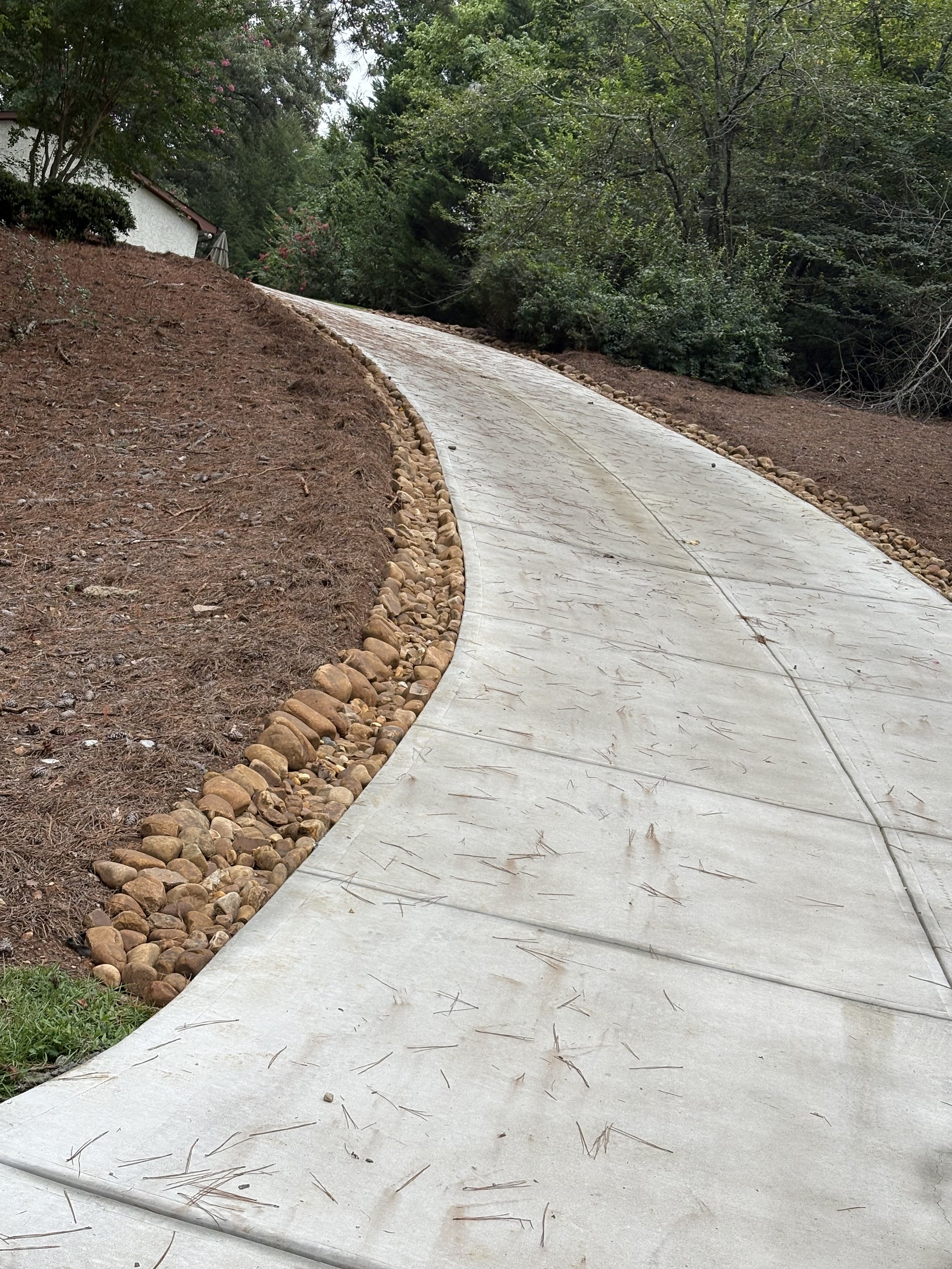 Curving concrete pathway lined with small rocks, on a slope with dirt on one side and trees on the other.