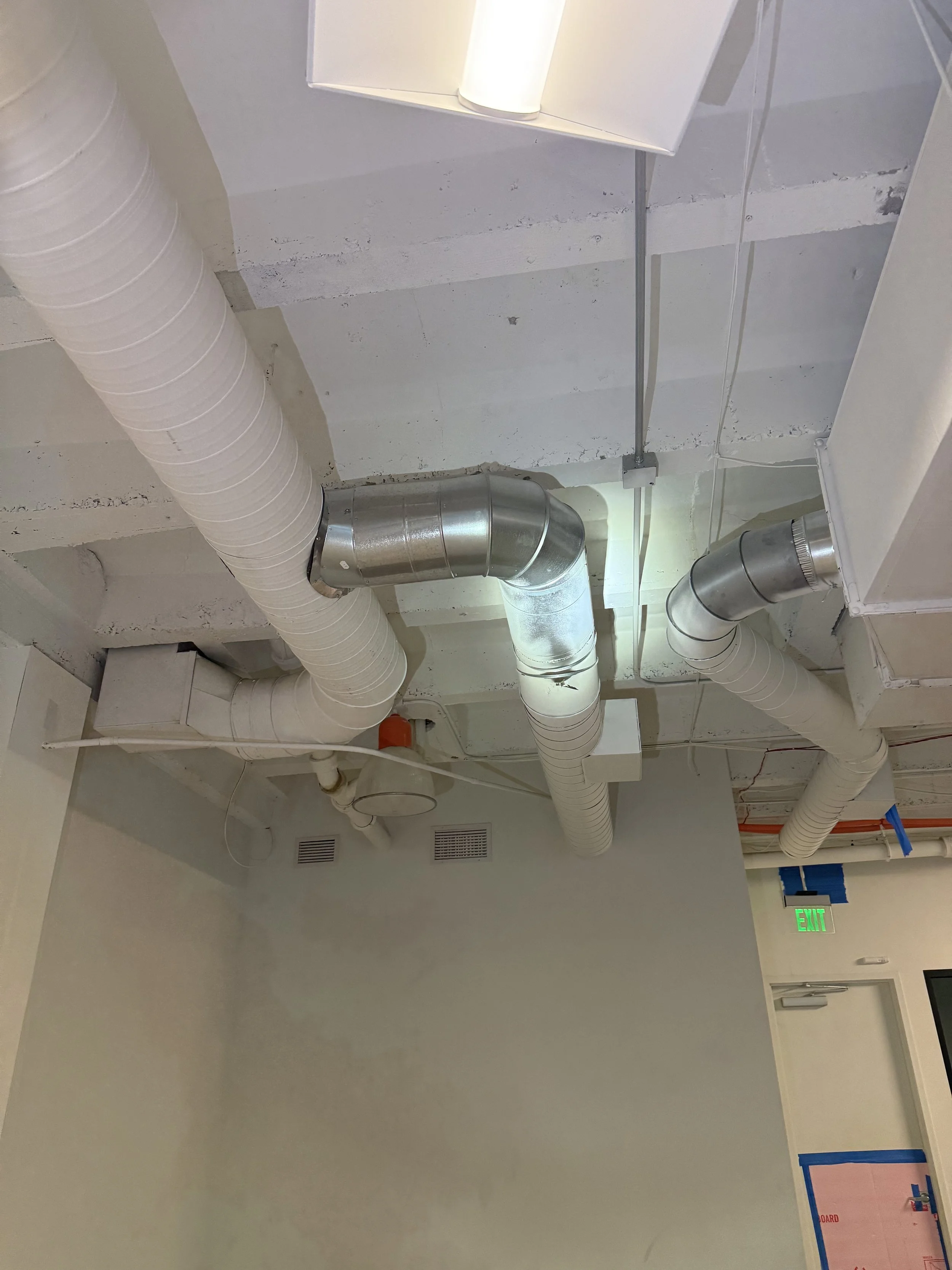 Ceiling with white ventilation ducts and a fluorescent light fixture, with a wall and an exit sign below.