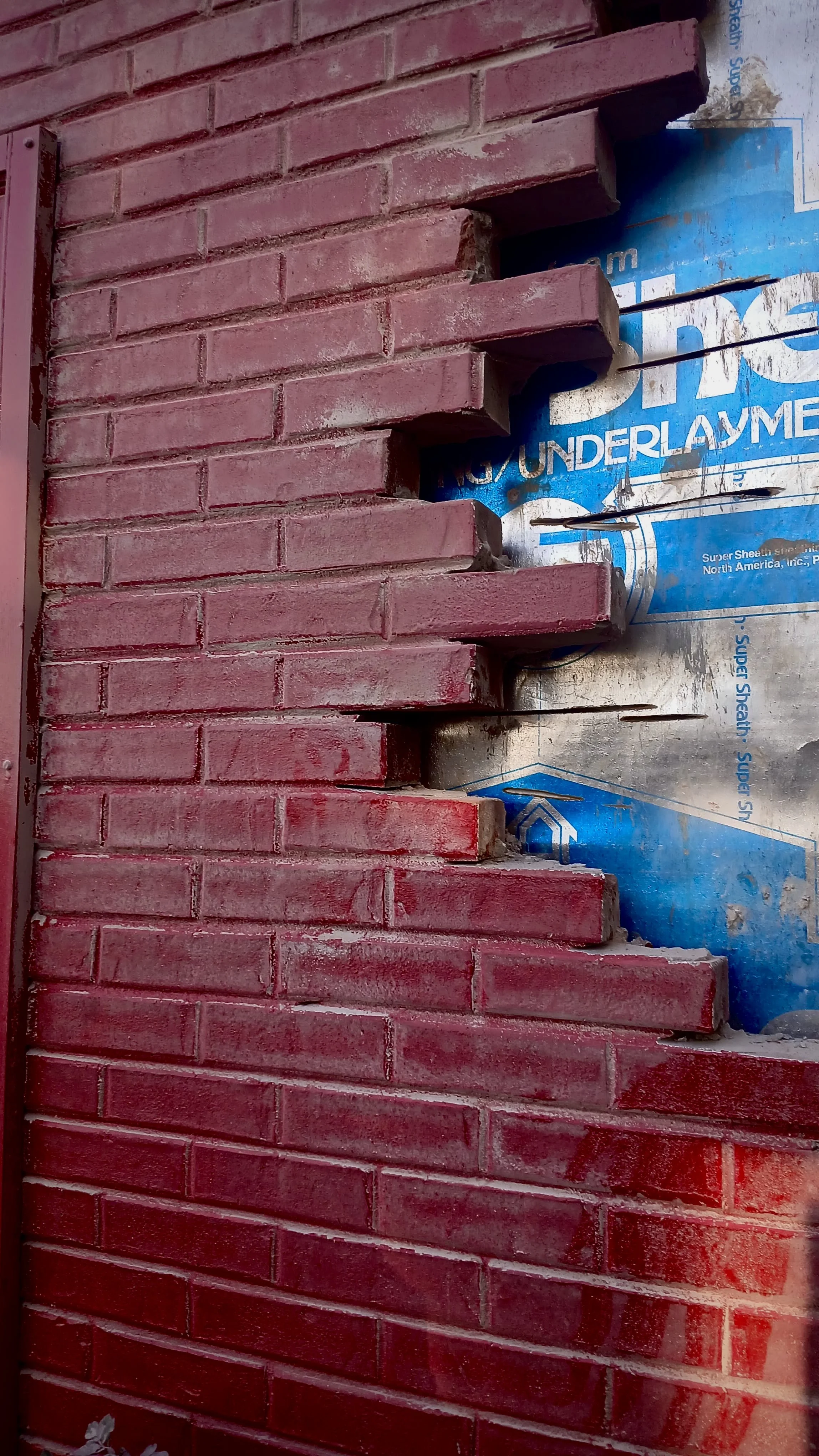 Close-up photo of a brick wall under construction with horizontal red bricks and metal support layer visible behind.