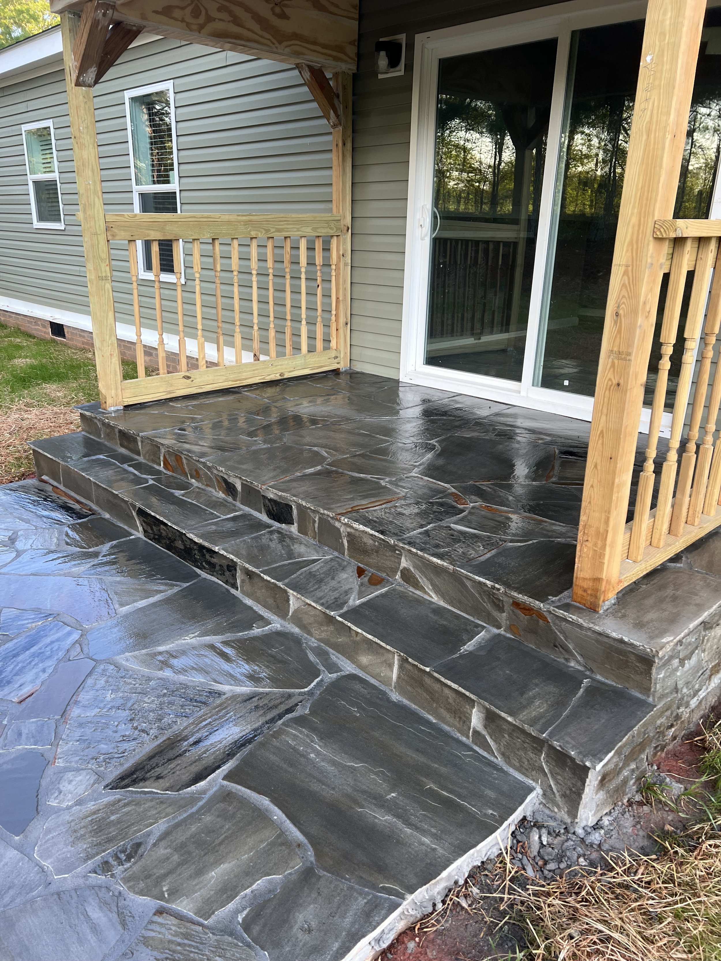 A newly constructed stone patio with steps leading to a sliding glass door on a house, with a small wooden railing on one side.