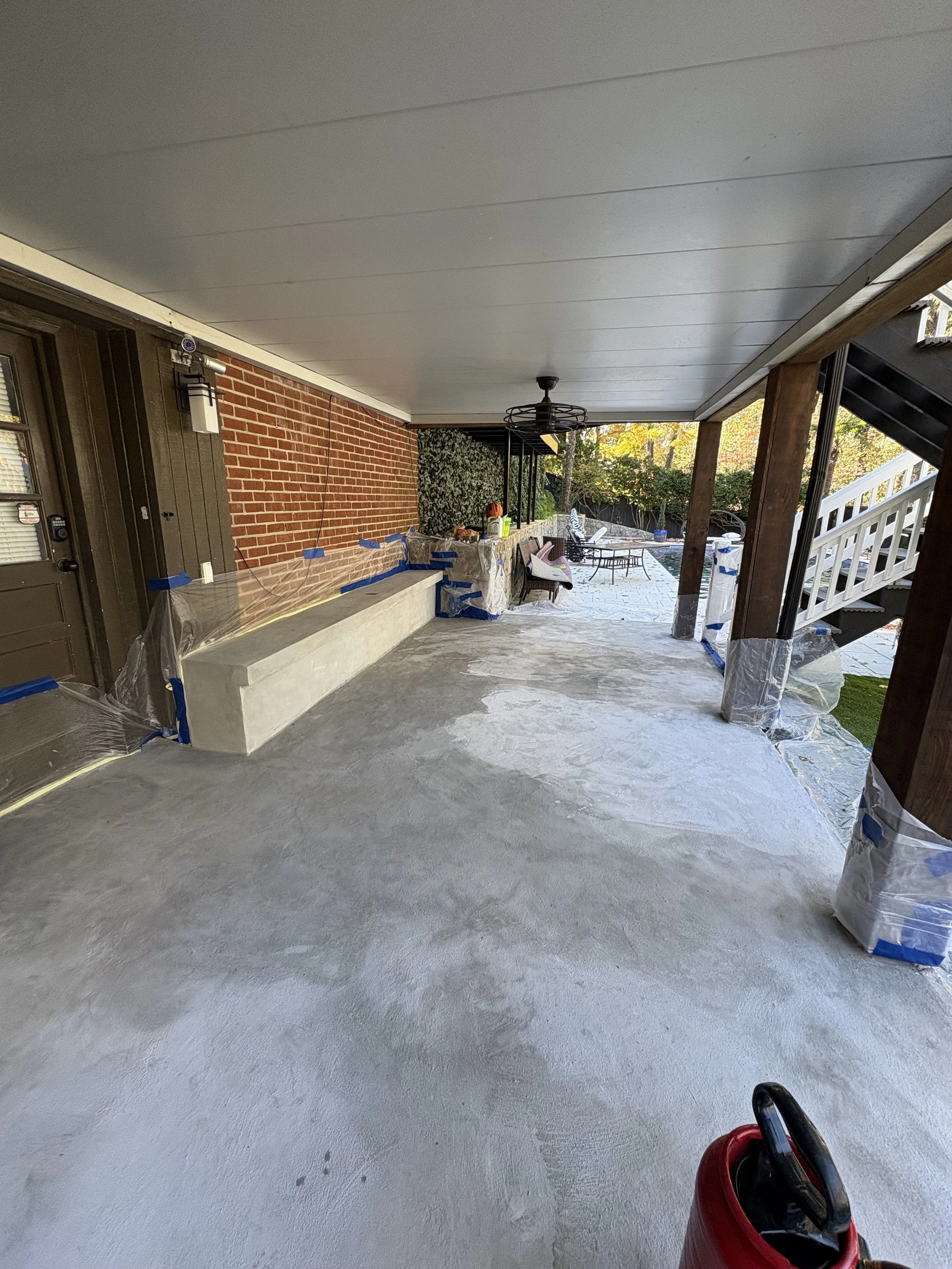 Covered porch area under renovation with concrete floor, brick wall, and outdoor furniture, with stairs leading to the yard and trees in the background.