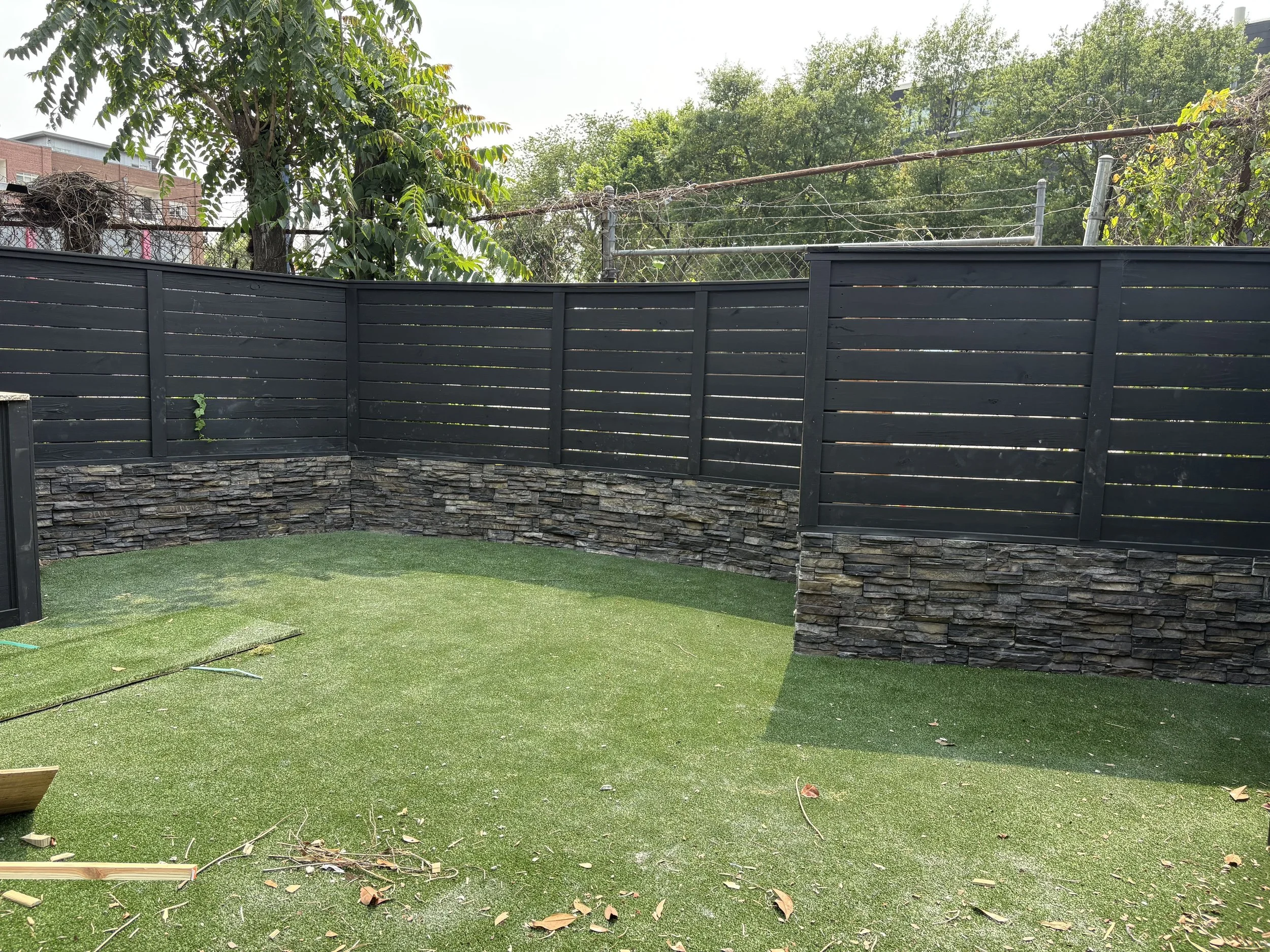 A backyard with green artificial grass, black wood fence with stone base, and trees in the background.