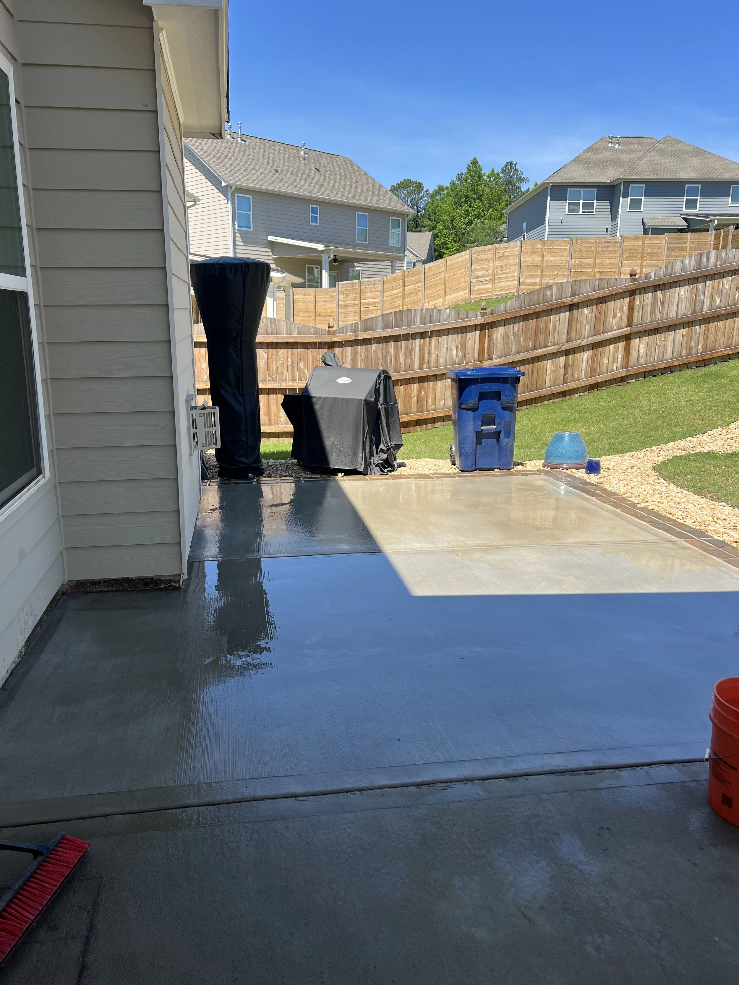 Recently cemented backyard patio with wet surface, outdoor furniture covered, trash bin, and garden decorations, with neighboring houses and wooden fence in background.