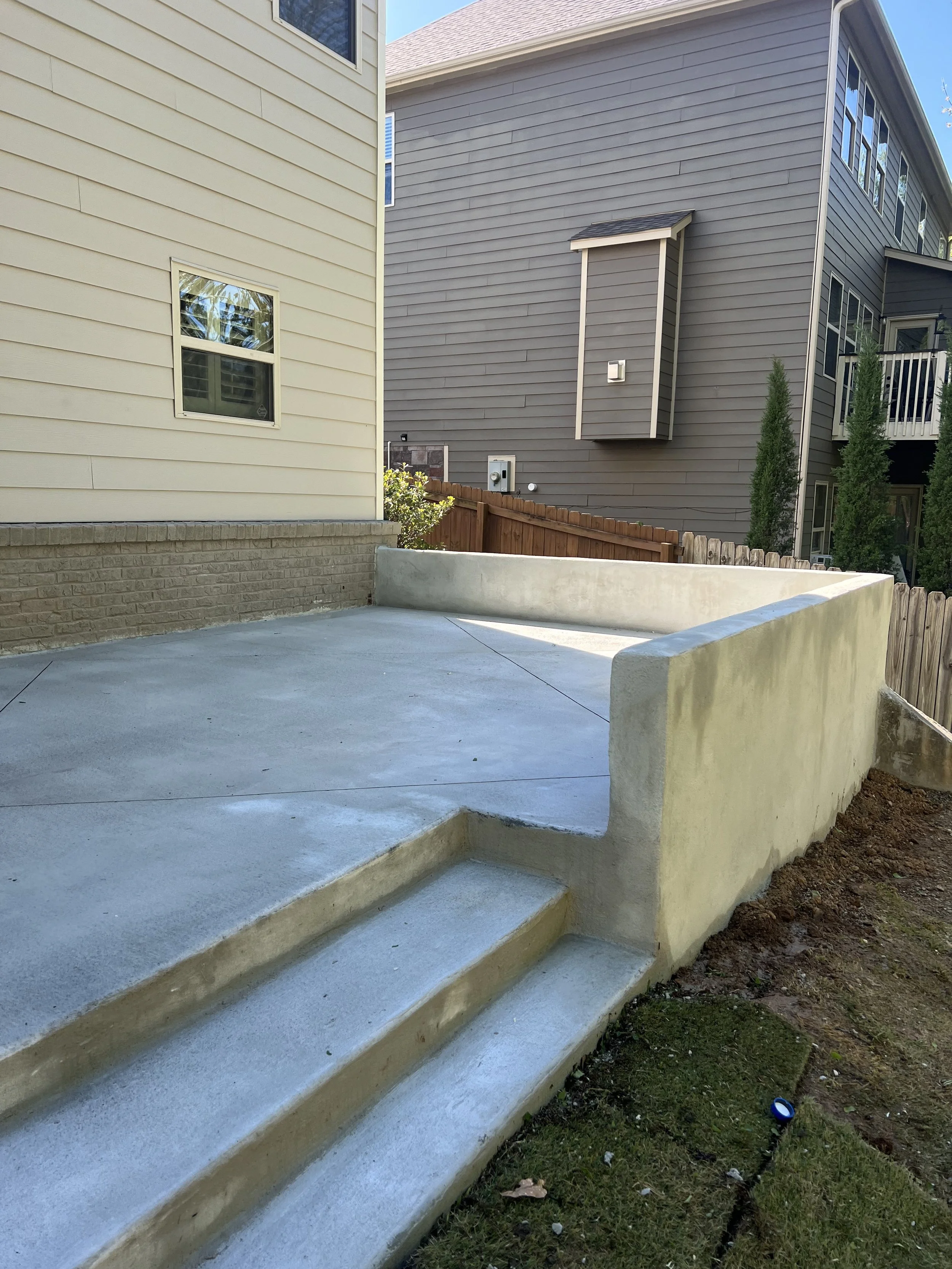 A newly constructed concrete patio with stairs leading up to it, situated behind a house, with neighboring houses and a wooden fence in the background.