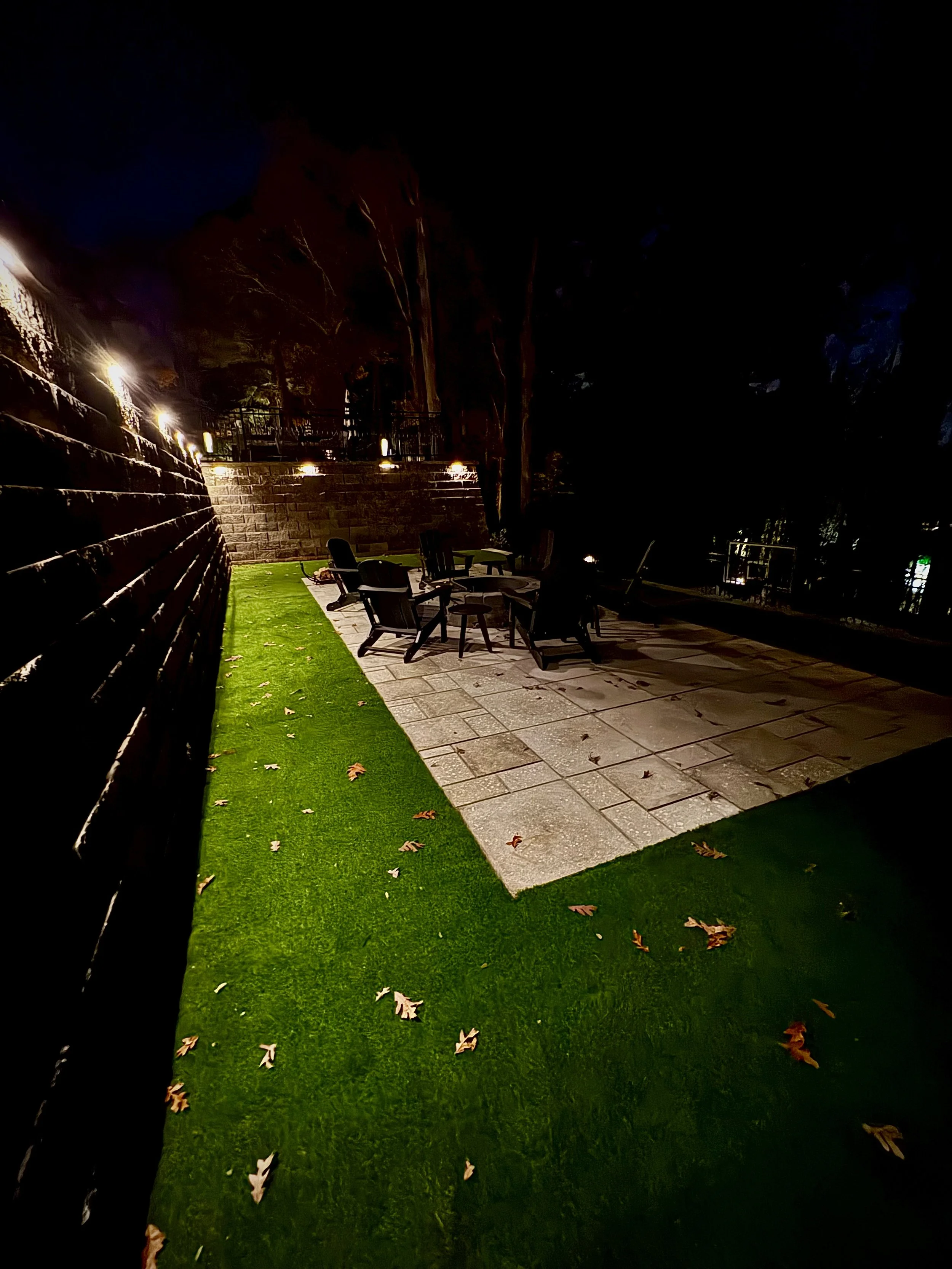 Night view of an outdoor seating area with a stone patio, black chairs, and a grassy strip, illuminated by lights along a wooden fence, with trees in the background.