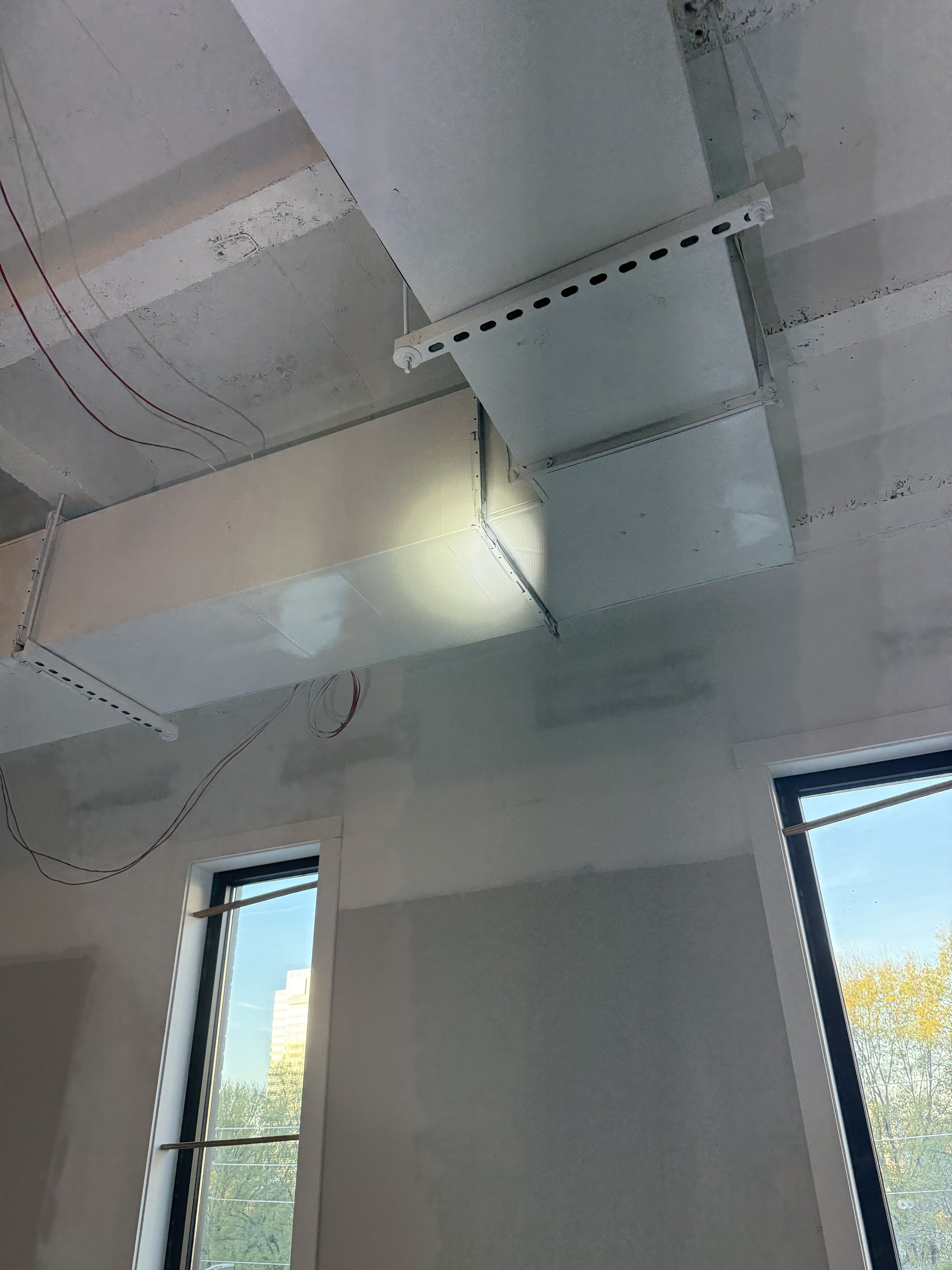 View of a ceiling with HVAC ductwork, exposed ceiling beams, and unfinished drywall in a room with large windows.