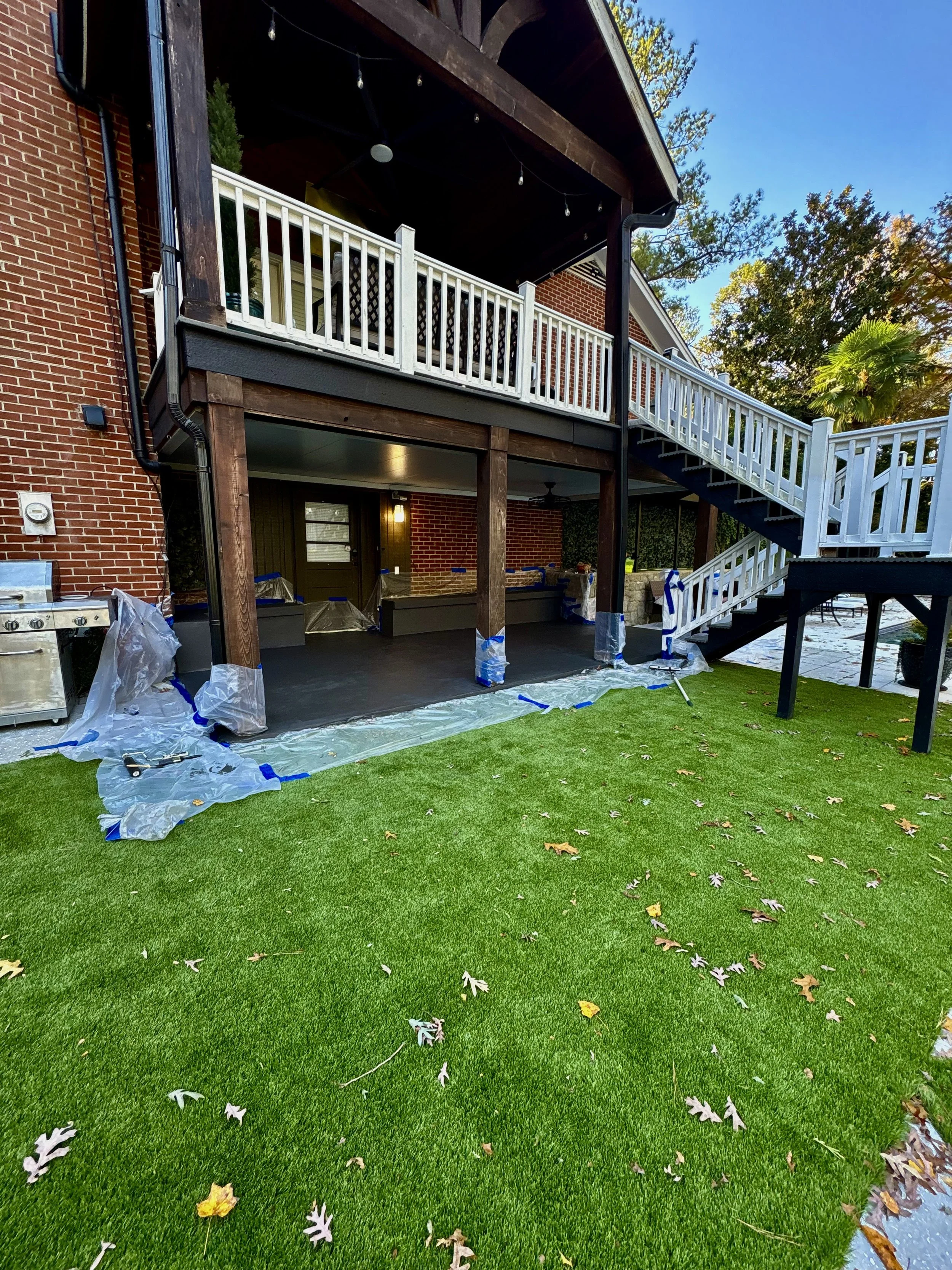 Backyard patio under construction with new flooring and a two-story wooden deck. The lower area is covered with a dark flooring, and the upper deck has white railings. Plastic protective covers are around the columns. The yard has green grass with sc