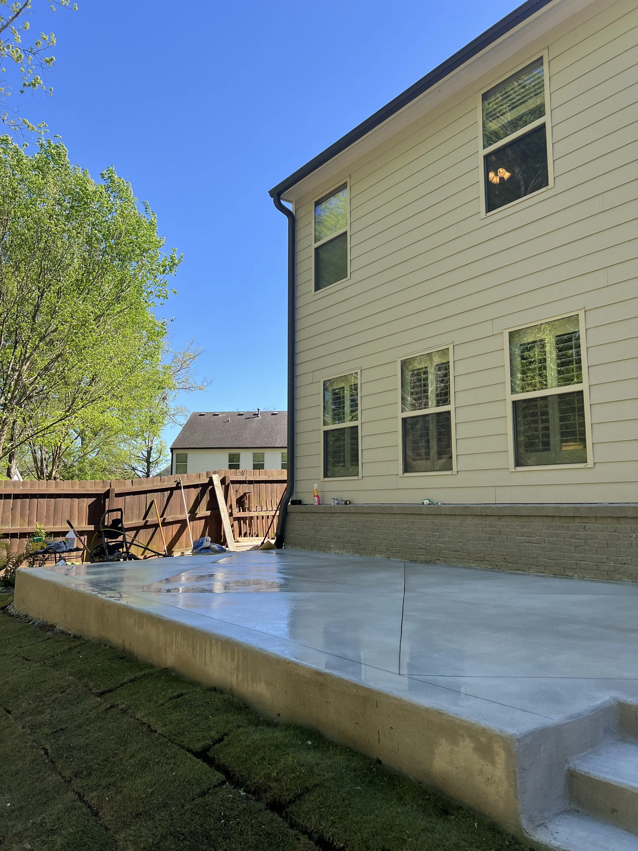 Newly poured concrete patio in backyard of a house with beige siding and multiple windows, fenced yard, and green trees.