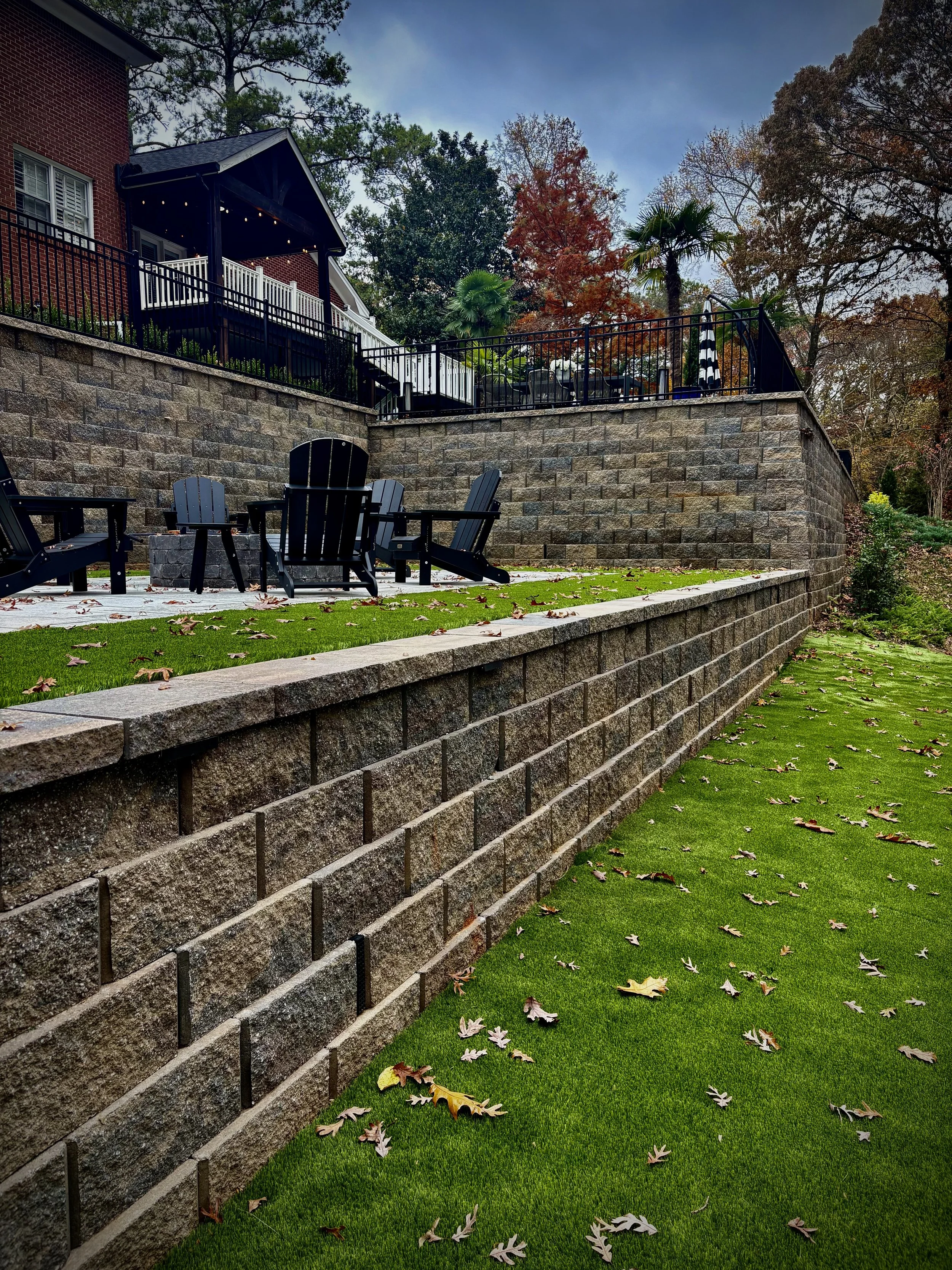 Backyard with a multi-level stone retaining wall, green grass, and outdoor seating area with black Adirondack chairs.
