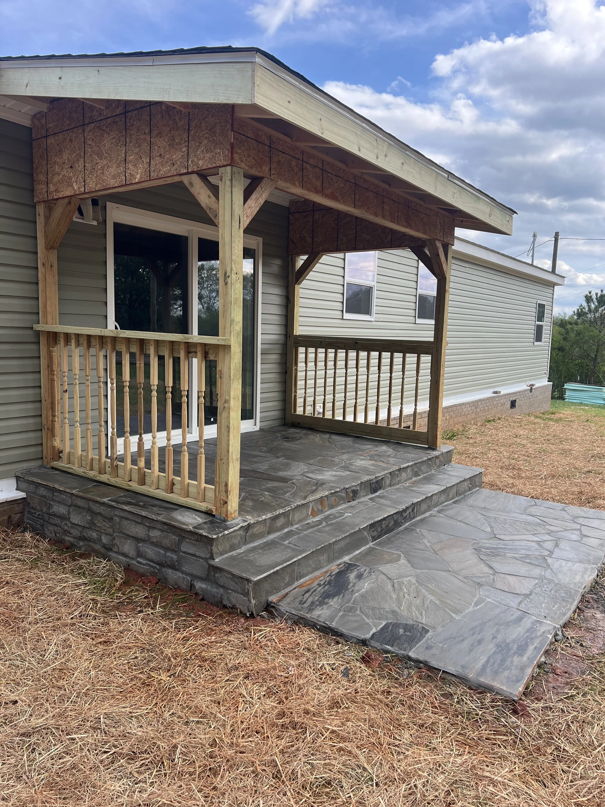 Newly built small front porch with stone steps and railing on a house with beige siding. The porch has a wooden frame and a roof, and the ground is covered with pine straw and gravel.