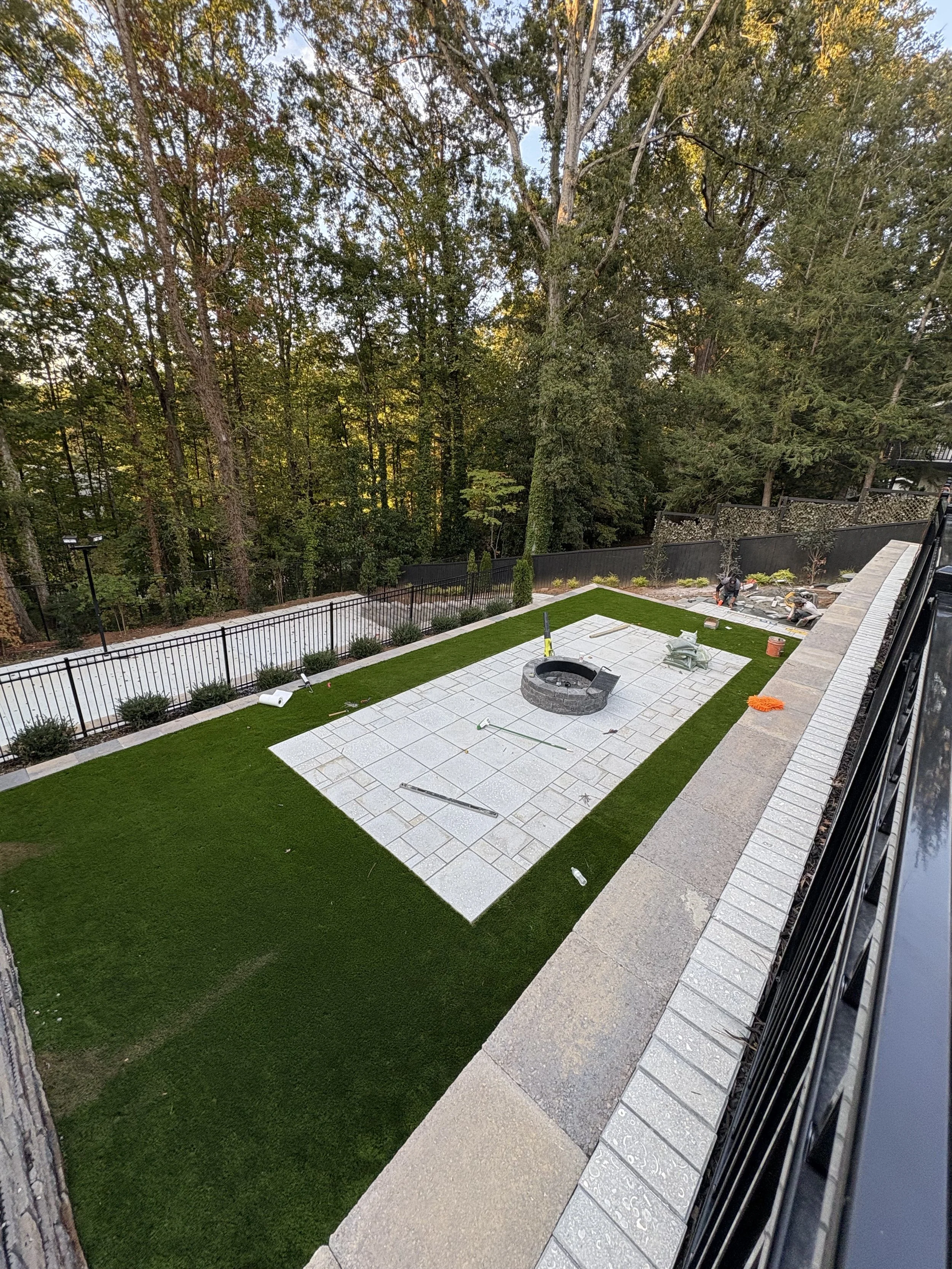 Backyard landscaping with newly installed paver patio, fire pit, artificial grass border, and ongoing construction work
