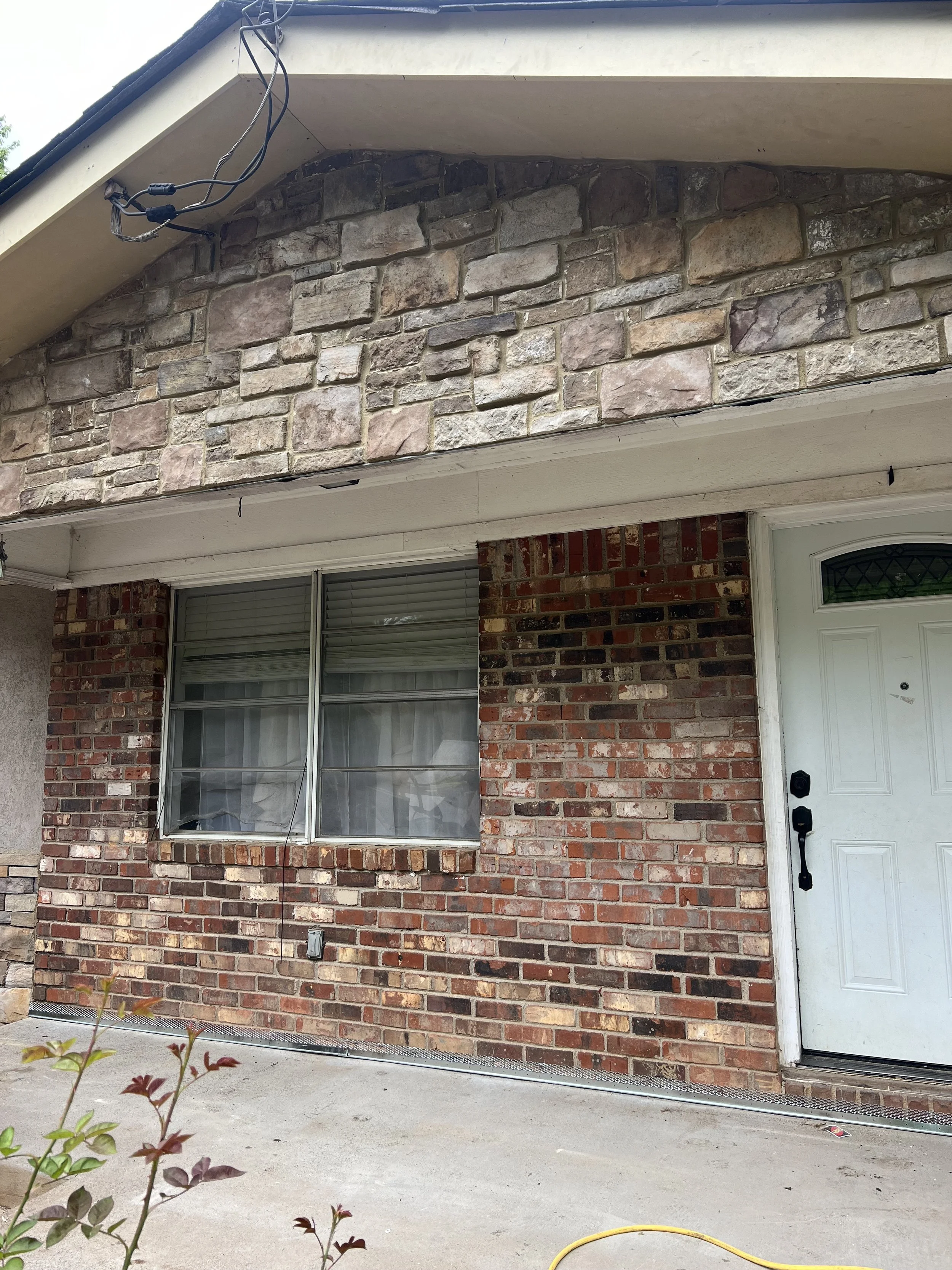 Close-up view of the front of a house showing brick exterior walls, a window with closed blinds, and a white door with a black handle and lock. Part of the roof and an open electrical conduit are visible.