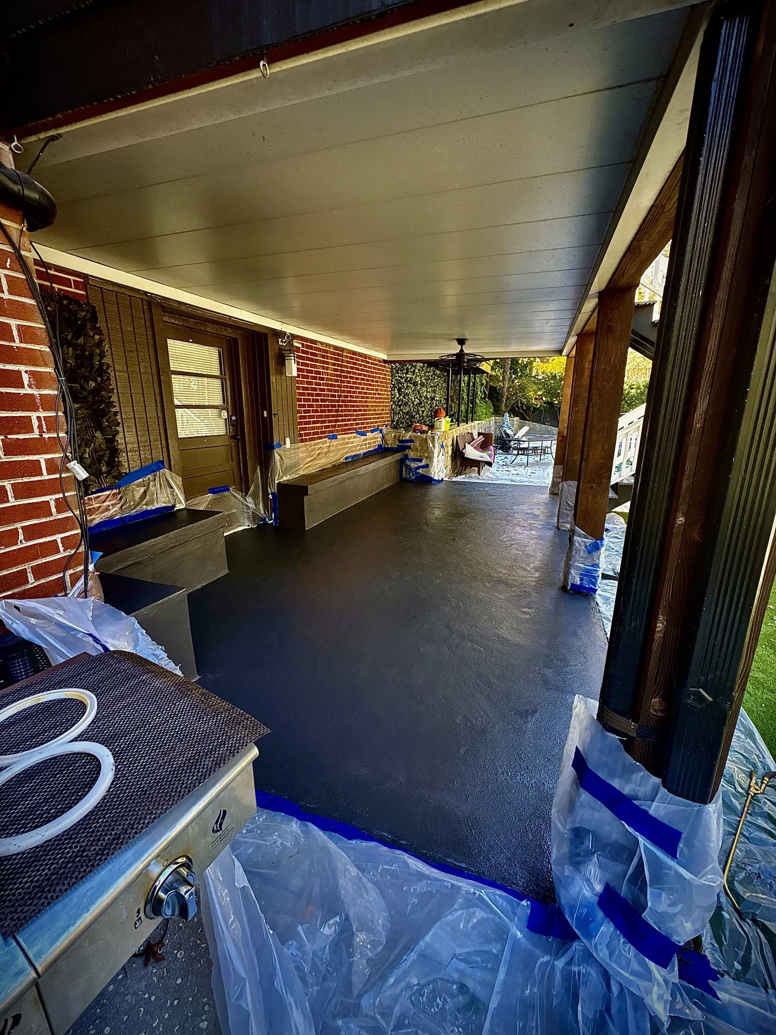 Covered porch area with newly painted black floor, plastic covers on furniture and edges, and partially painted brick wall in the background.