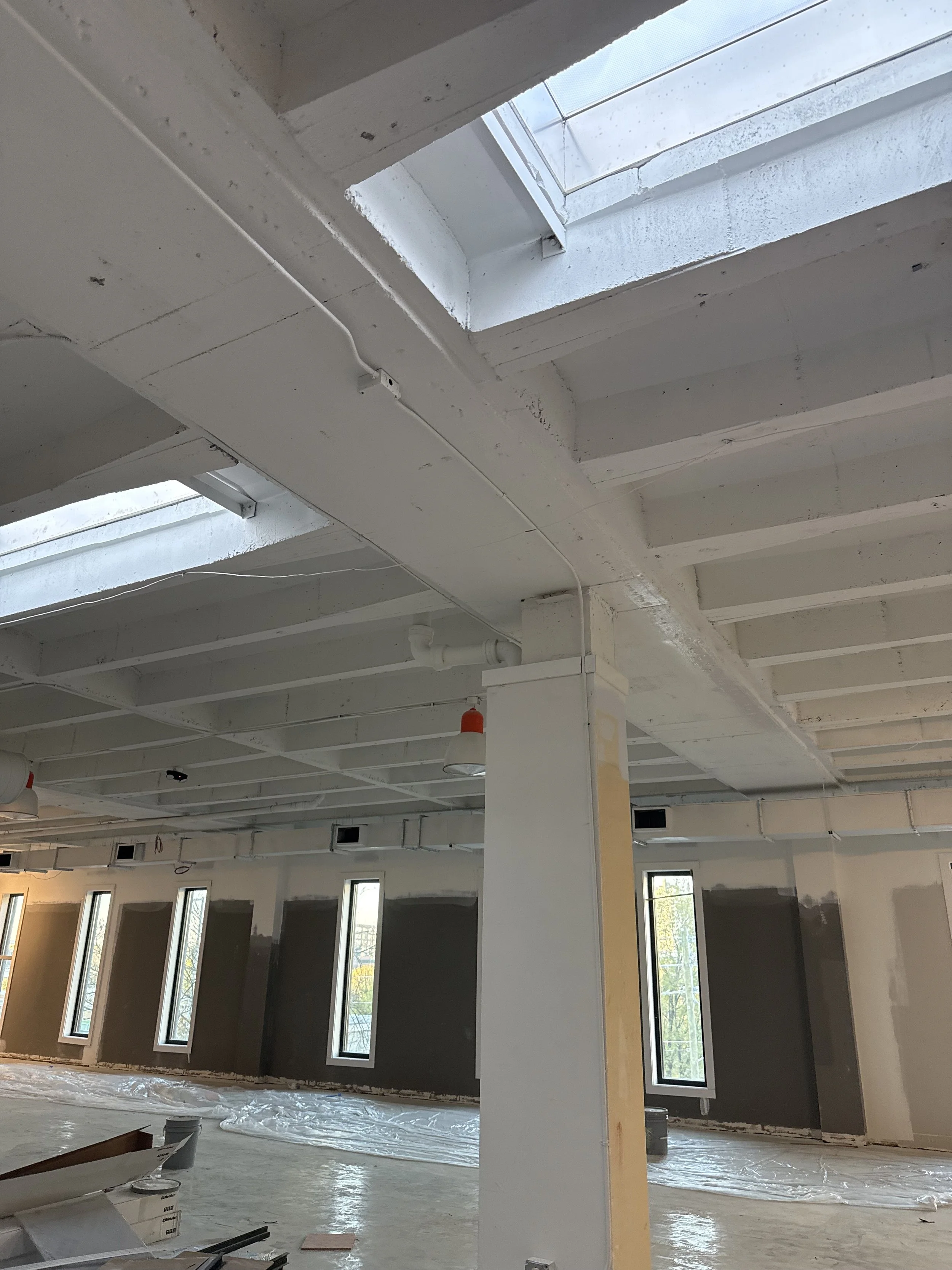 Interior view of a building under construction with exposed white ceiling beams, skylights, and tall narrow windows along the walls. Construction materials and plastic sheeting are on the unfinished floor.