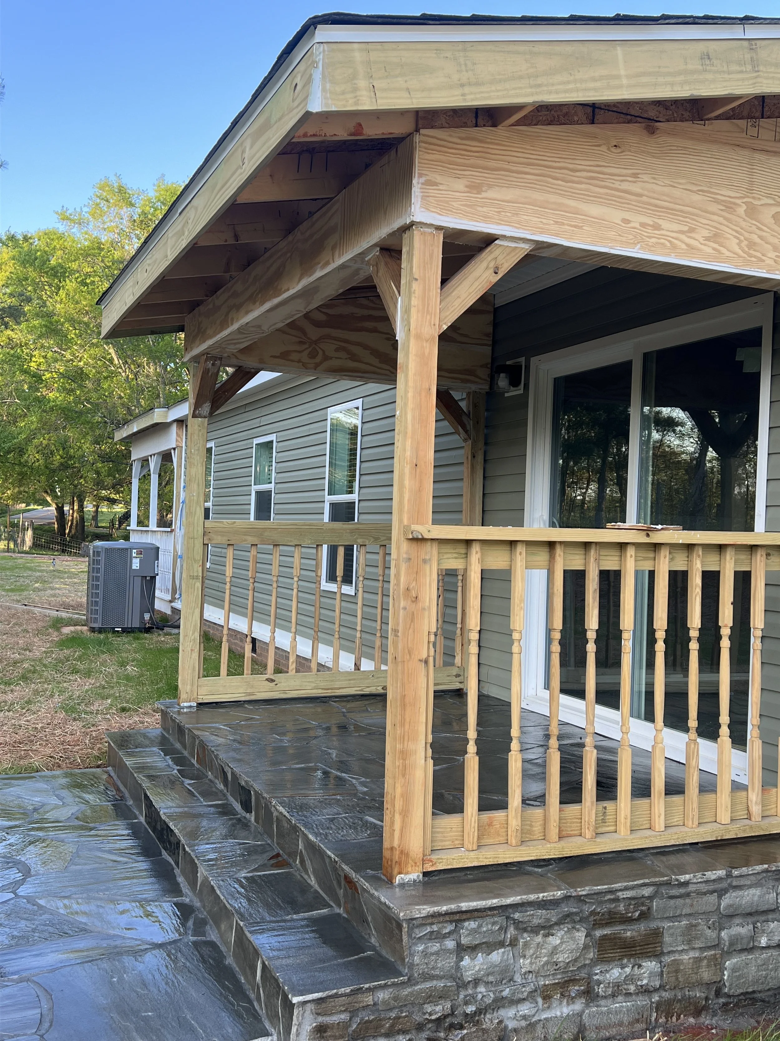 Newly built wooden deck and porch attached to a house, with steps leading up, finished with stone and dark tiles, and a view of a backyard with trees.
