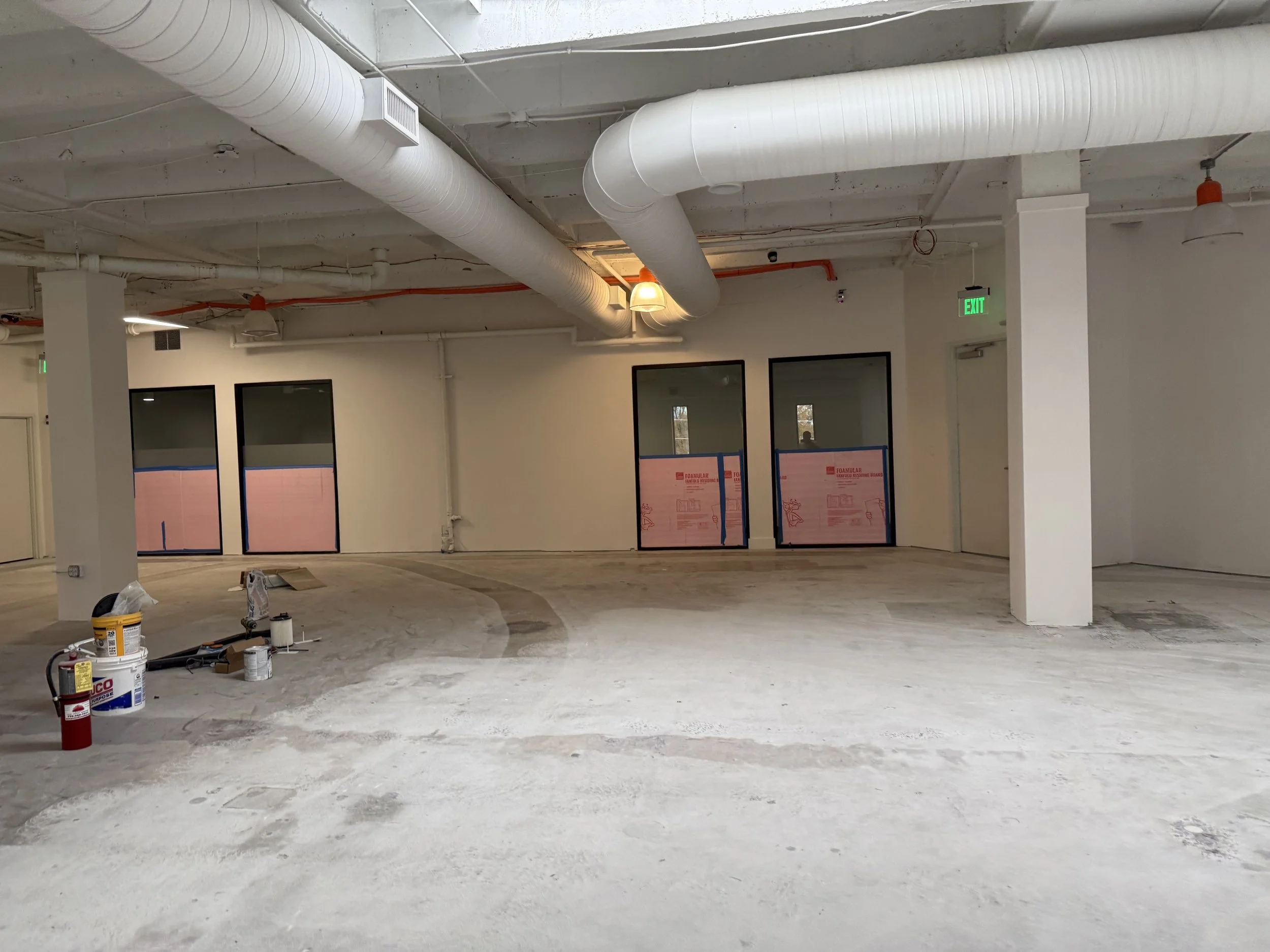 Interior of a building under construction with exposed ceiling ductwork, construction materials on the floor, and windows with construction covering.
