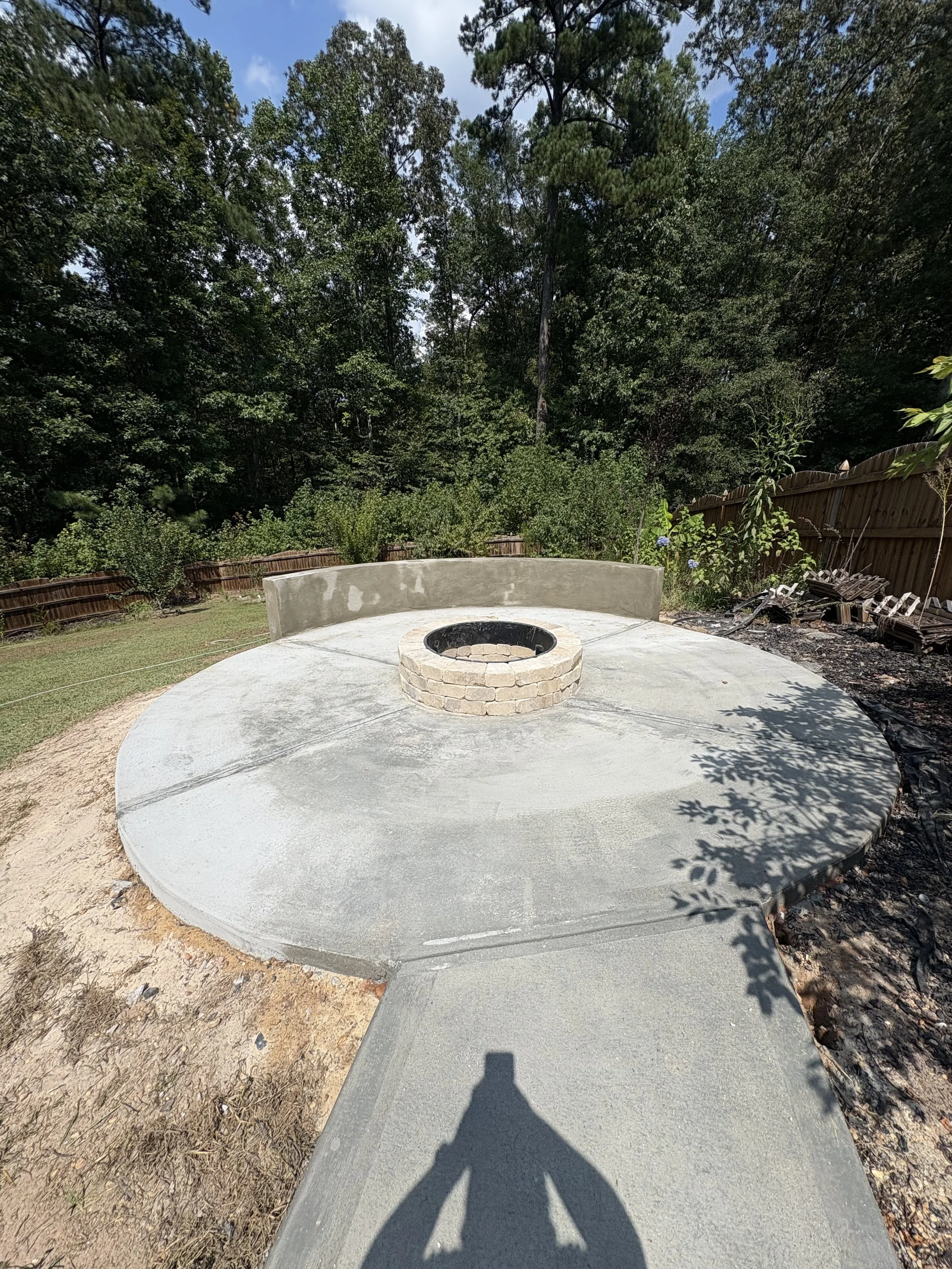 A circular concrete patio with a fire pit in the center, surrounded by a wooded backyard with a fence, trees, and plants.
