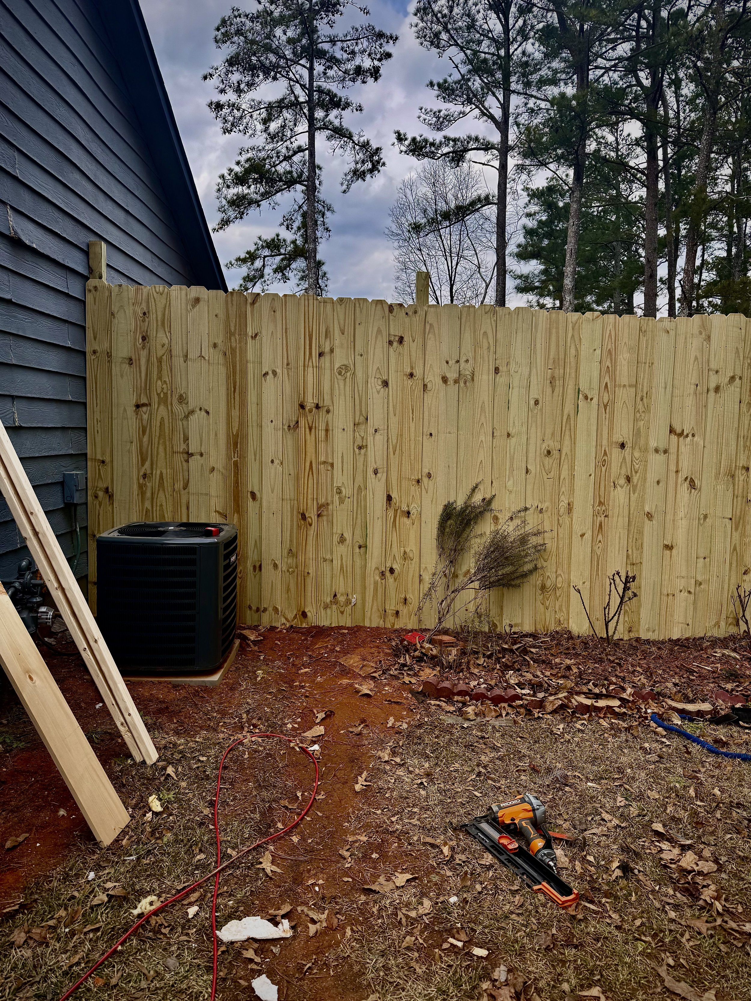 A backyard with a new wooden privacy fence under construction, with trees in the background and construction tools on the ground.