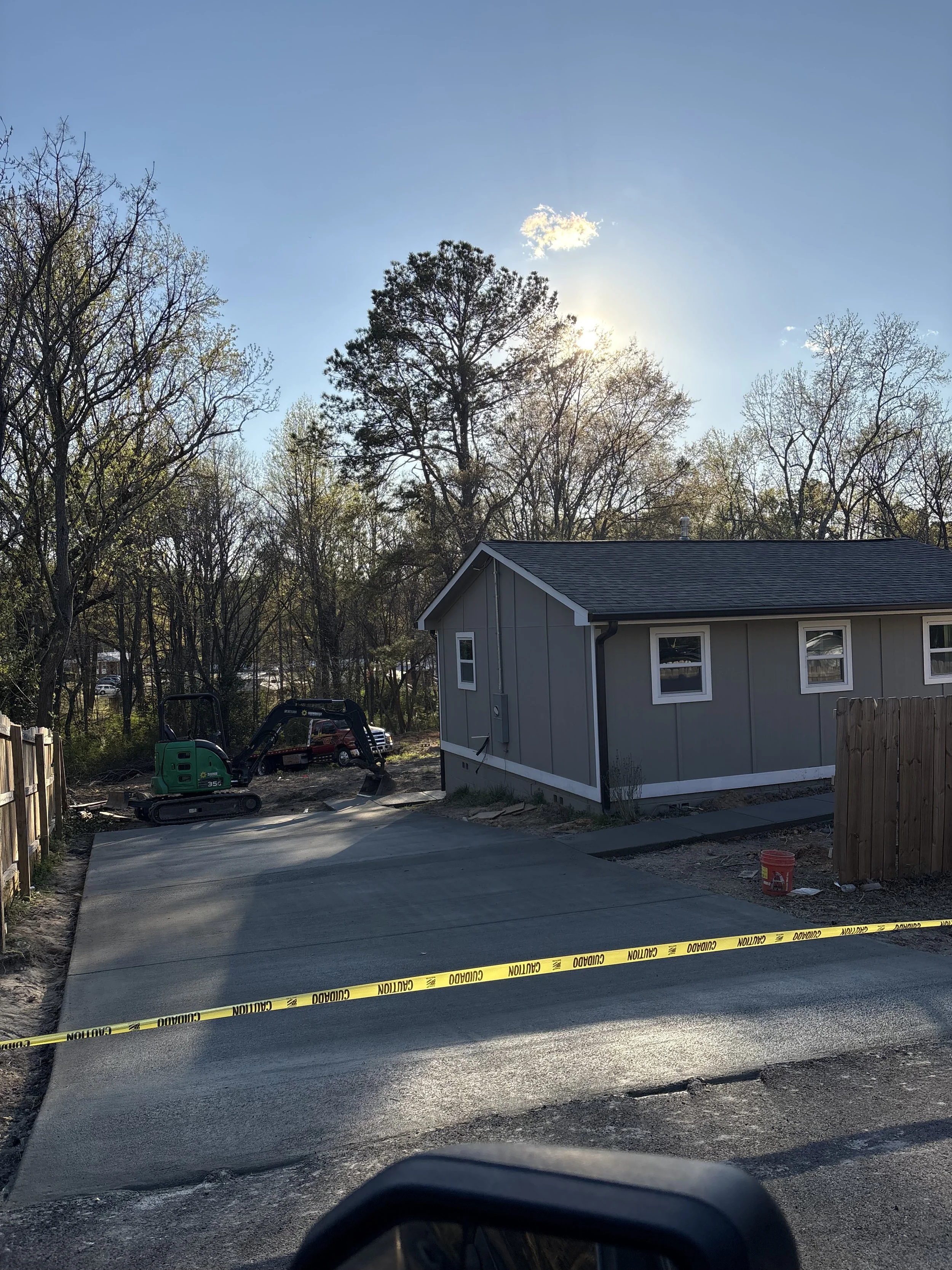 Newly paved concrete driveway in front of a gray house, with construction equipment and caution tape, and trees in the background with sunlight shining through.