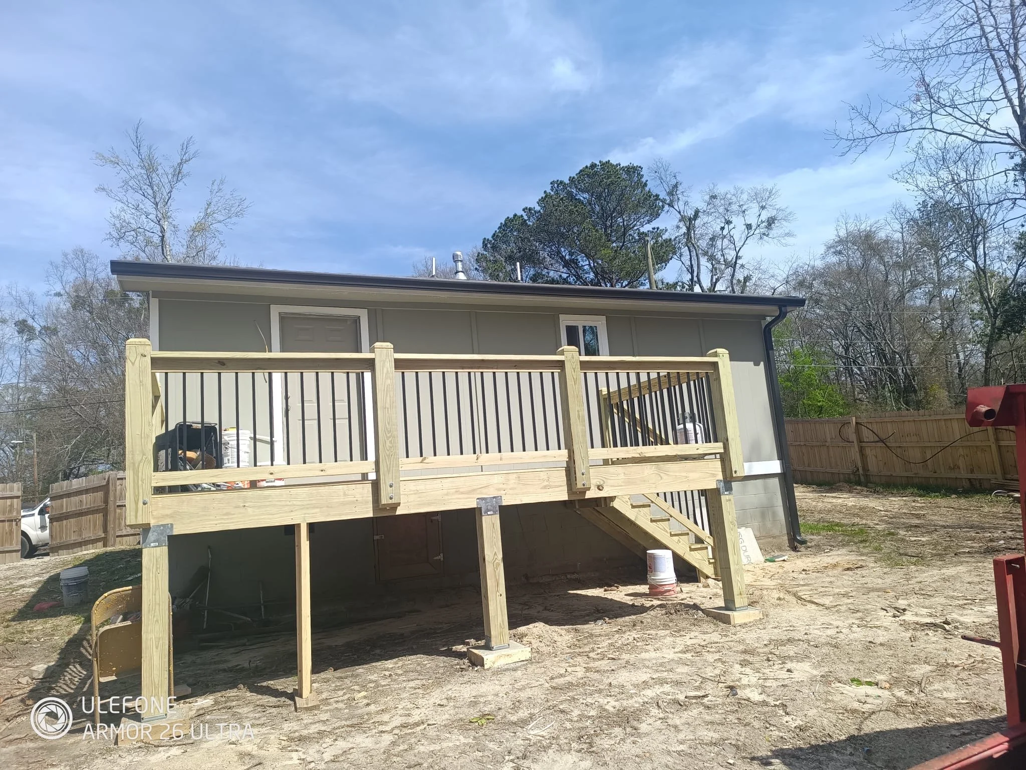 New wooden deck being built on the back of a house with outdoor stairs and a railing, on a dirt backyard with a fence and trees.