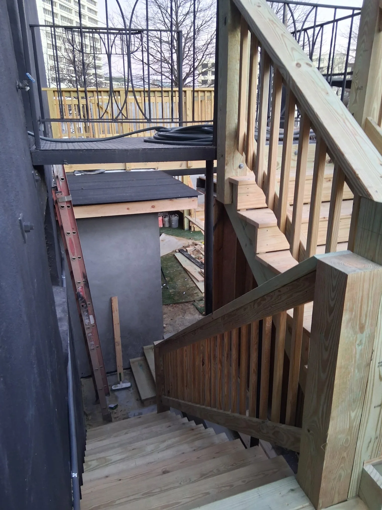 Photo of a wooden staircase with railing under construction, with tools, a ladder, and construction materials visible. In the background, a fenced outdoor area with trees and buildings.