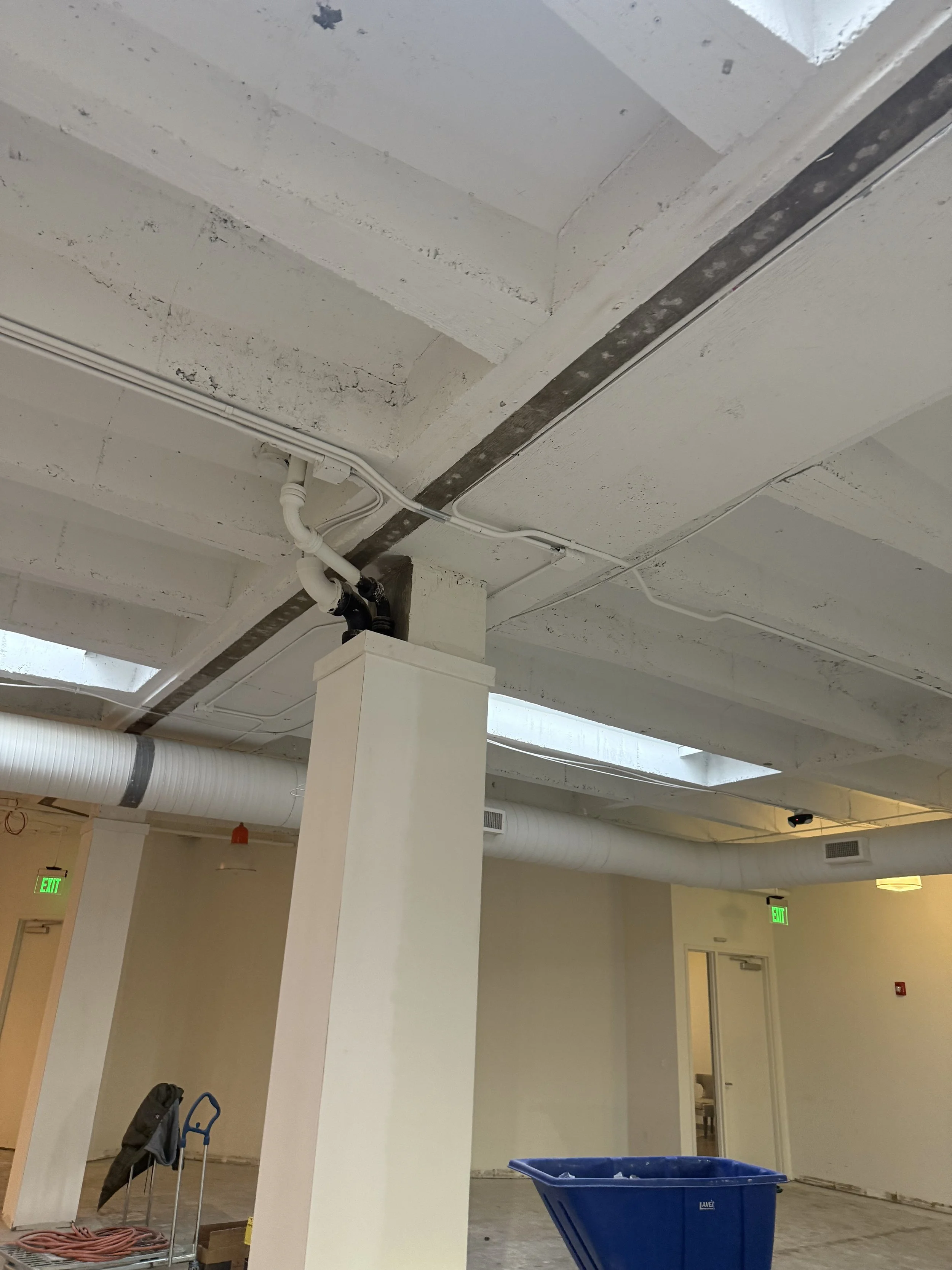 Interior view of a room under construction or renovation, showing exposed ceiling with pipes and wiring, a blue container, a black backpack on a chair, and open doors.
