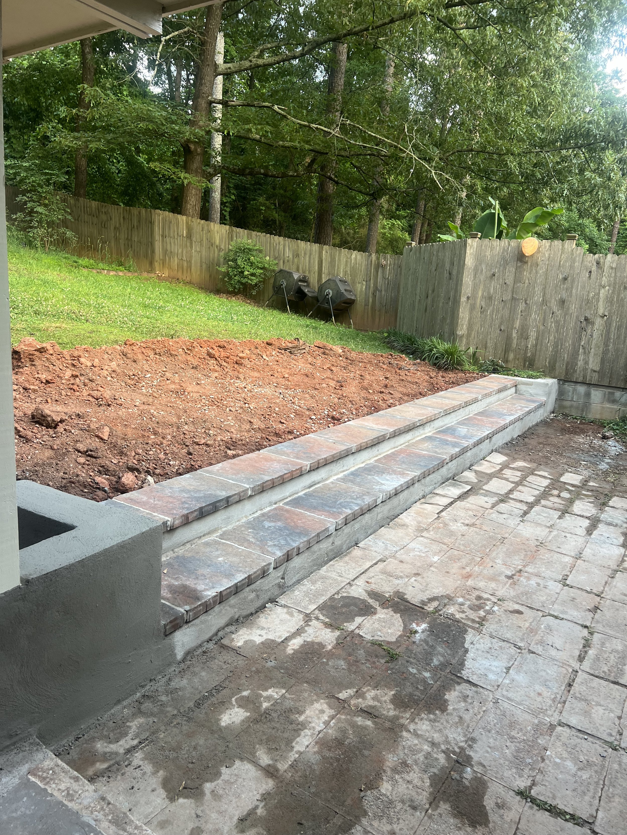 A backyard patio area under construction with stone and brick steps, grass and mulch beds, a fence, two black compost bins, and trees in the background.