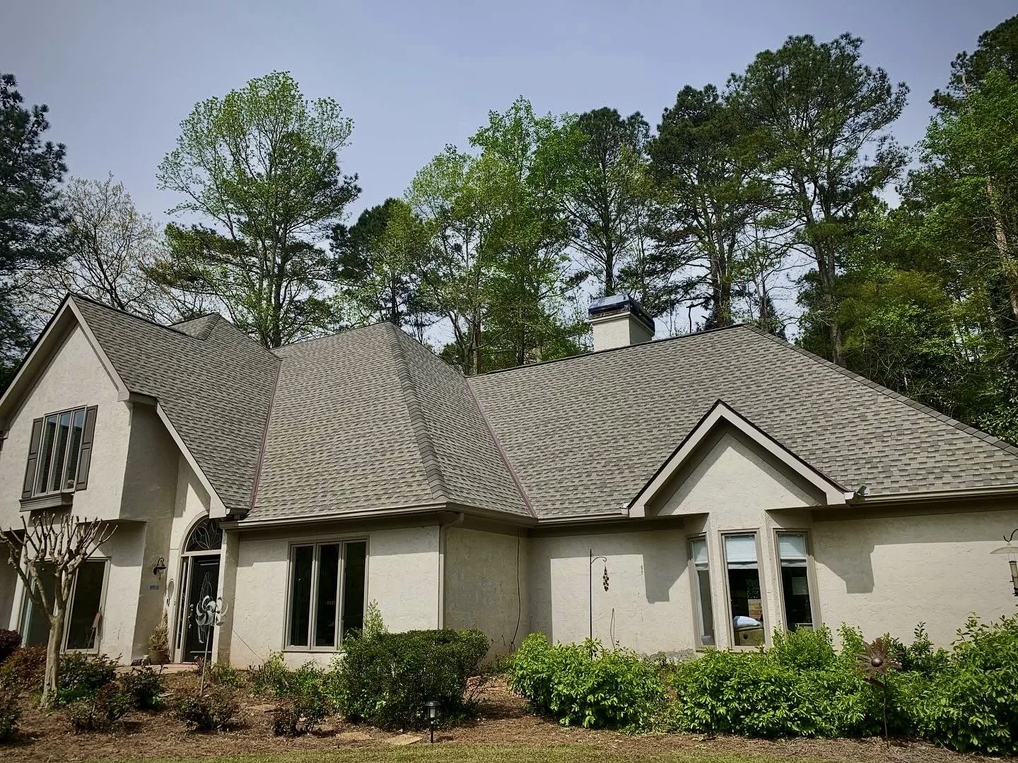 A house with a light-colored exterior and multiple gabled roofs, surrounded by green bushes and trees, with a background of tall trees under a clear sky.