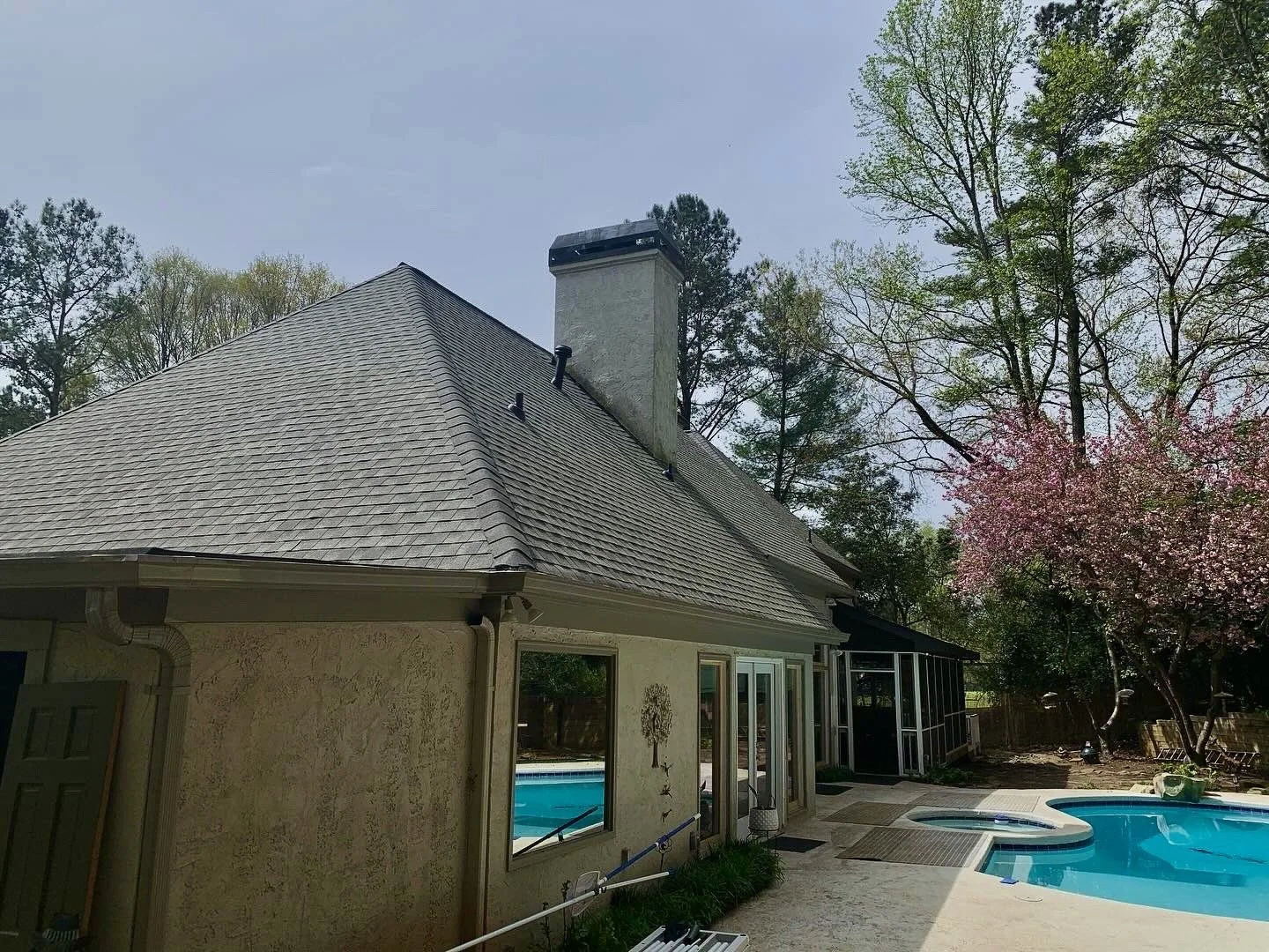 A backyard scene featuring a house with a pitched roof, a chimney, and a swimming pool. There are trees in the background, including one with pink blossoms.