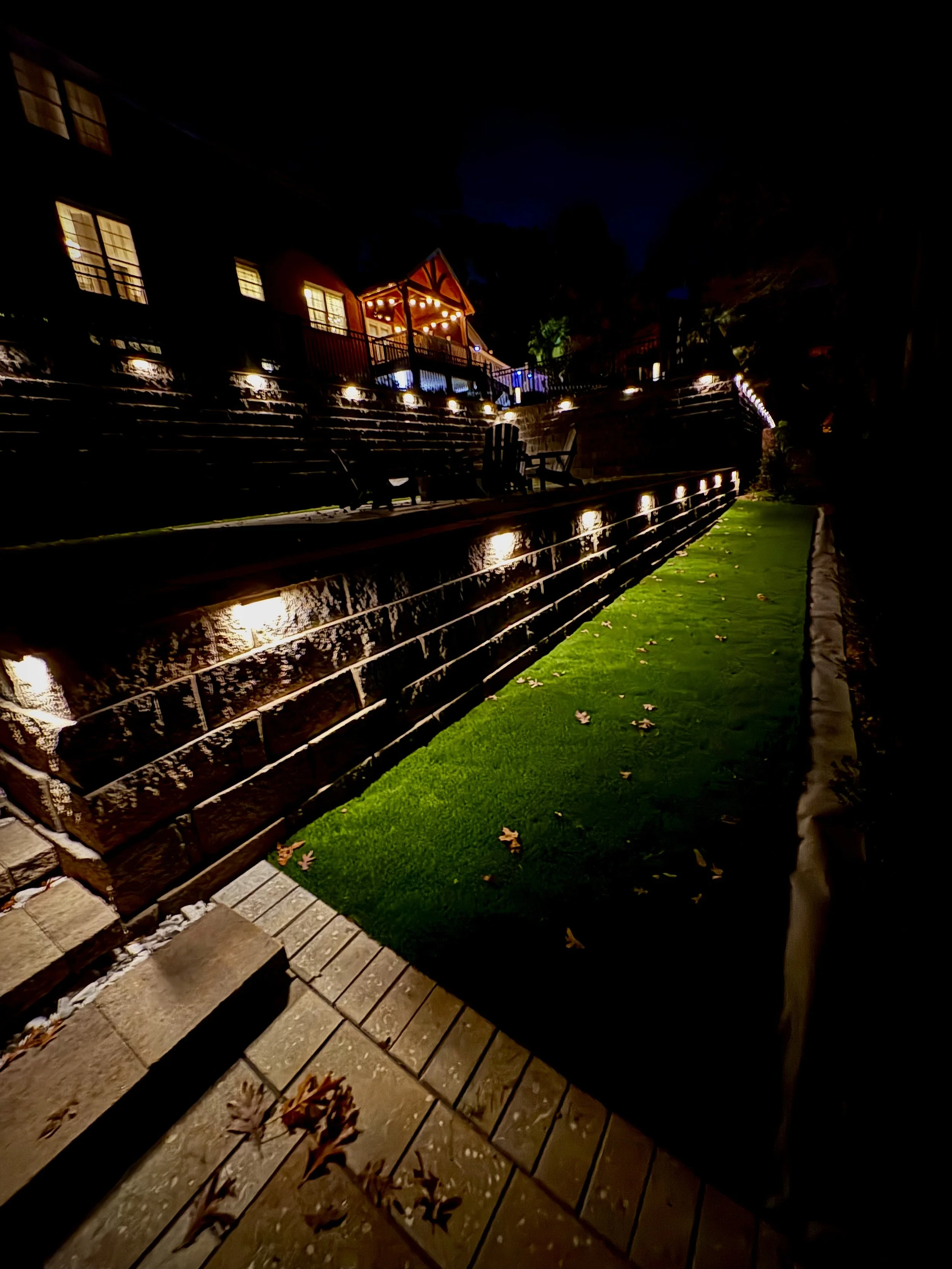 Night view of an outdoor terraced area with steps, illuminated pathway lights, a small patch of green grass, and a house or building with large windows and a porch with string lights.