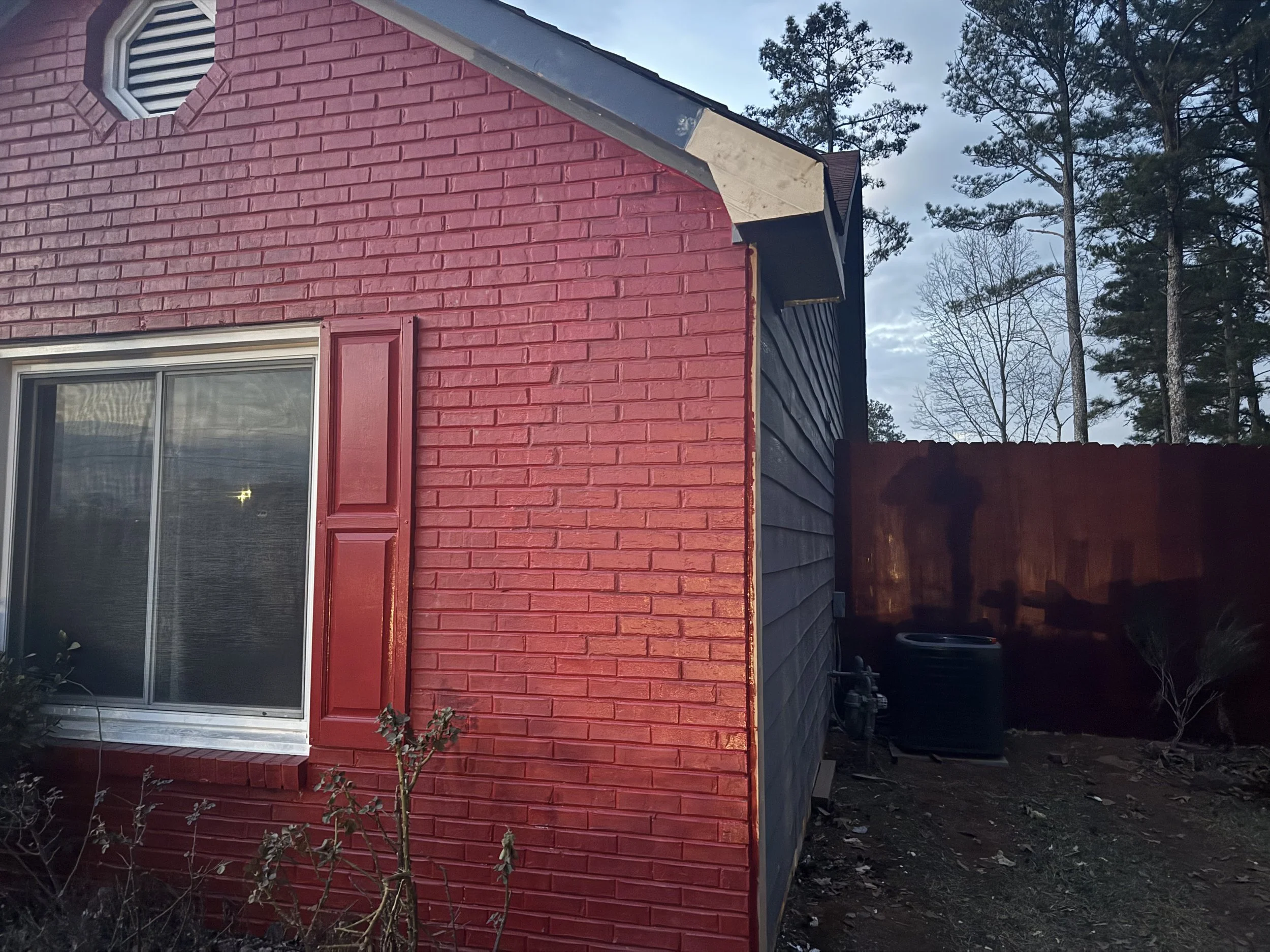 Close-up of a corner of a house with red brick exterior, black siding, and a window with closed blinds, with trees and a fence in the background.