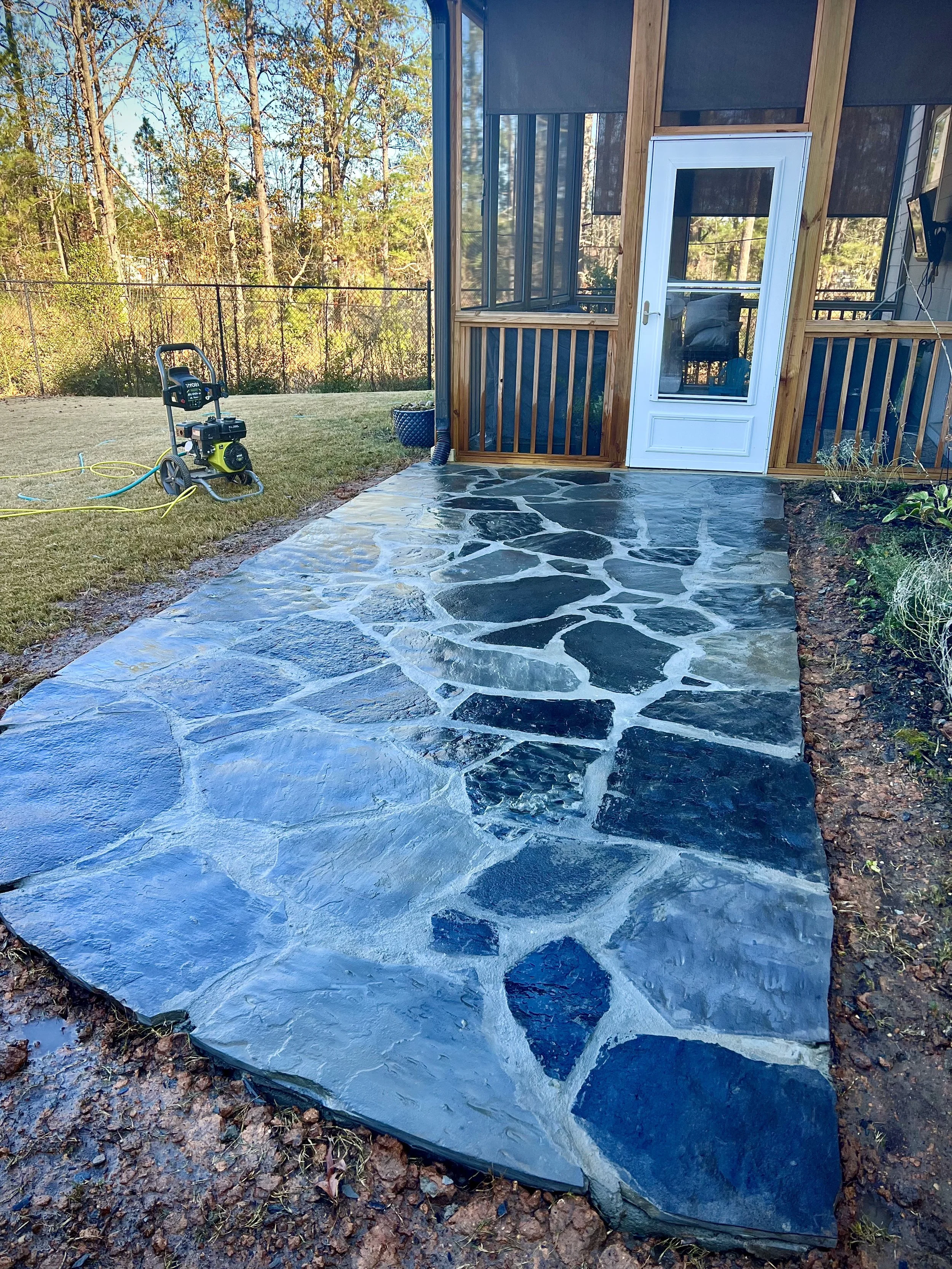 A newly installed stone patio leading to a back door of a wooden house with a screened porch. There is a lawn with gardening equipment nearby and a chain-link fence in the background.