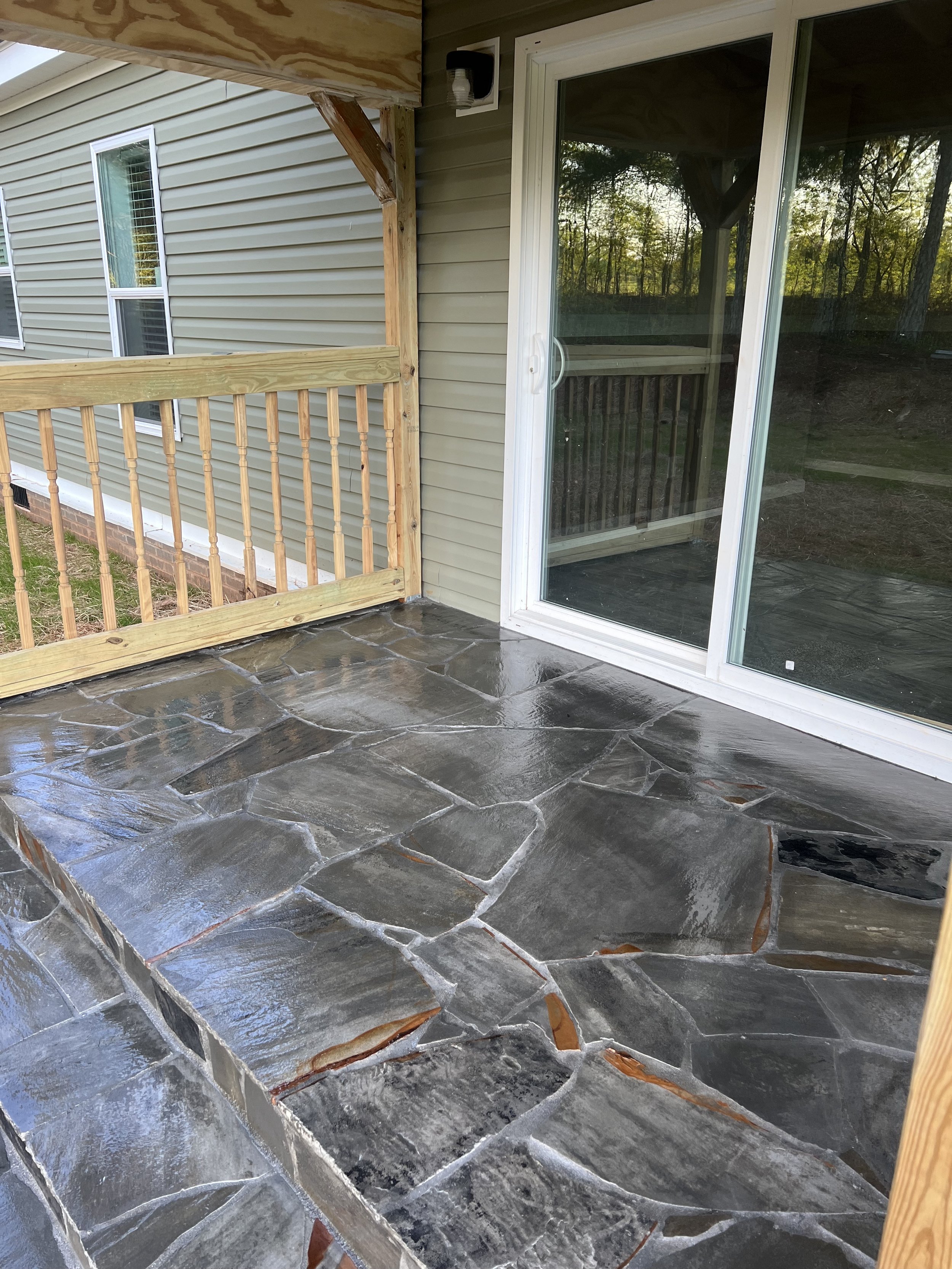 A small outdoor patio with a stone floor, a sliding glass door, and a wooden railing under construction. The patio is attached to a house with beige siding.