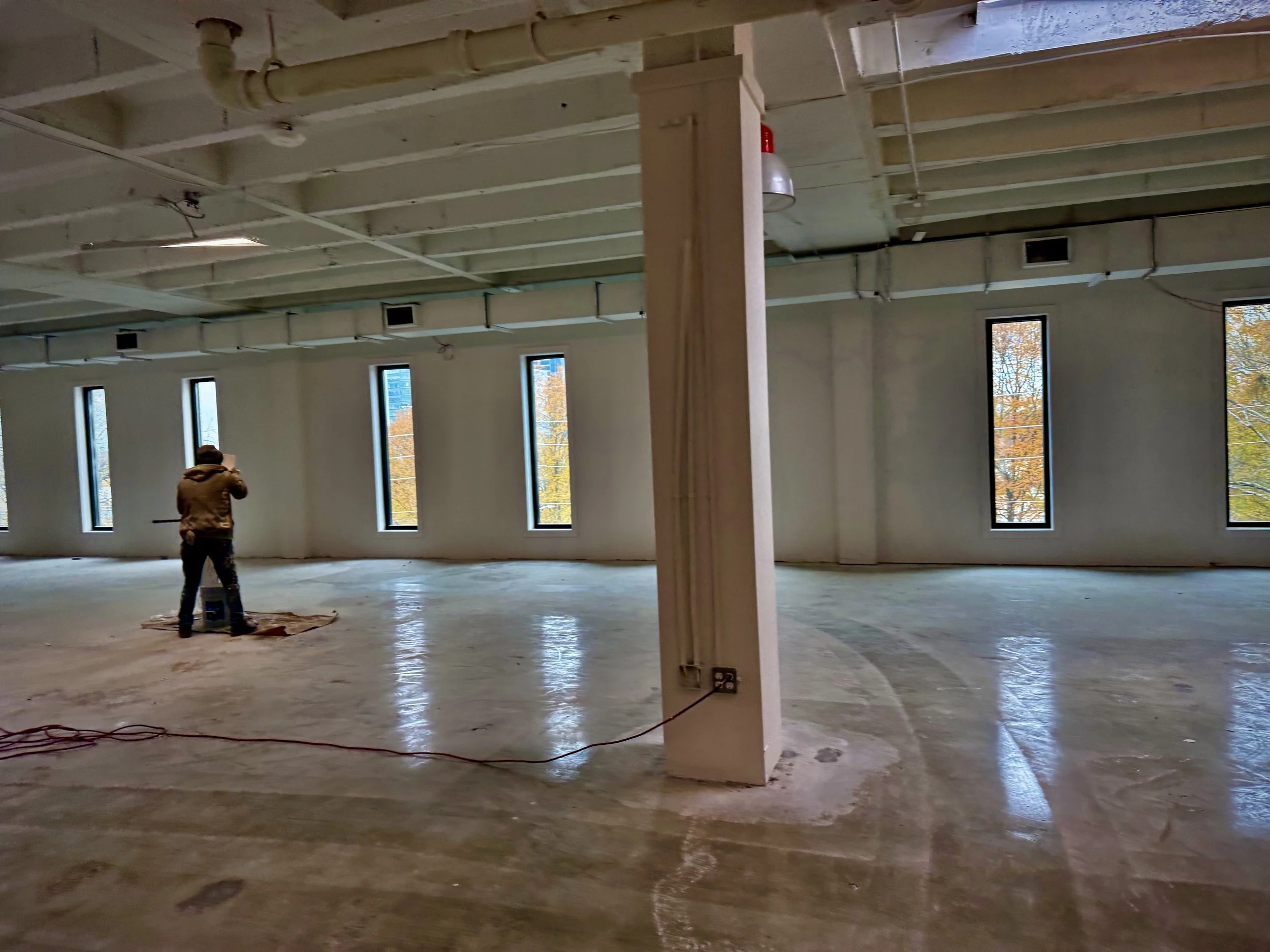 Interior of a building under construction with a worker standing on plastic tarp, a pillar in the foreground, and tall, narrow windows showing autumn trees outside.