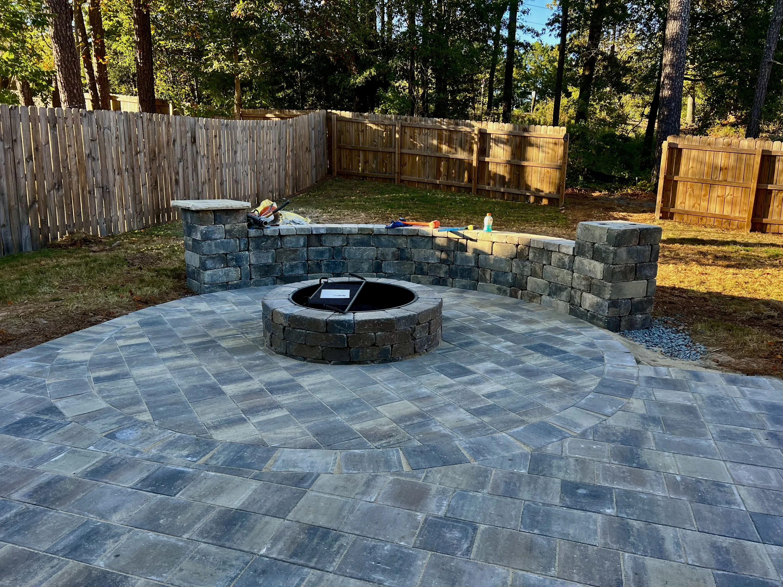 A backyard with a newly built stone patio, a fire pit, and a stone retaining wall, surrounded by wood fencing and tall trees.