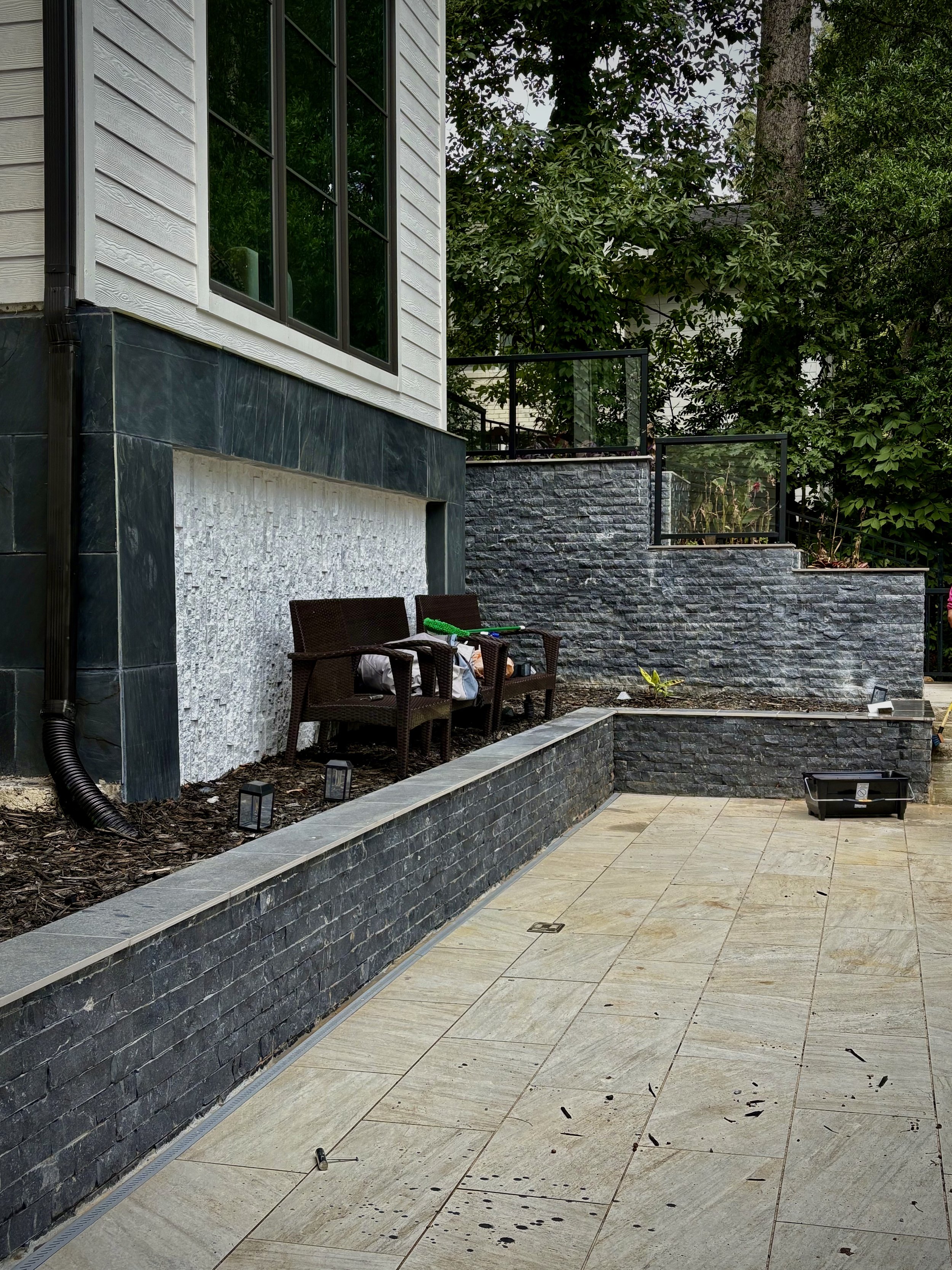 Outdoor patio with stone and tile surfaces, two brown wicker chairs with cushions, assorted items on the chairs, green garden tools, black planter box, surrounding trees and plantings, modern house with large window, black stone wall, and glass raili
