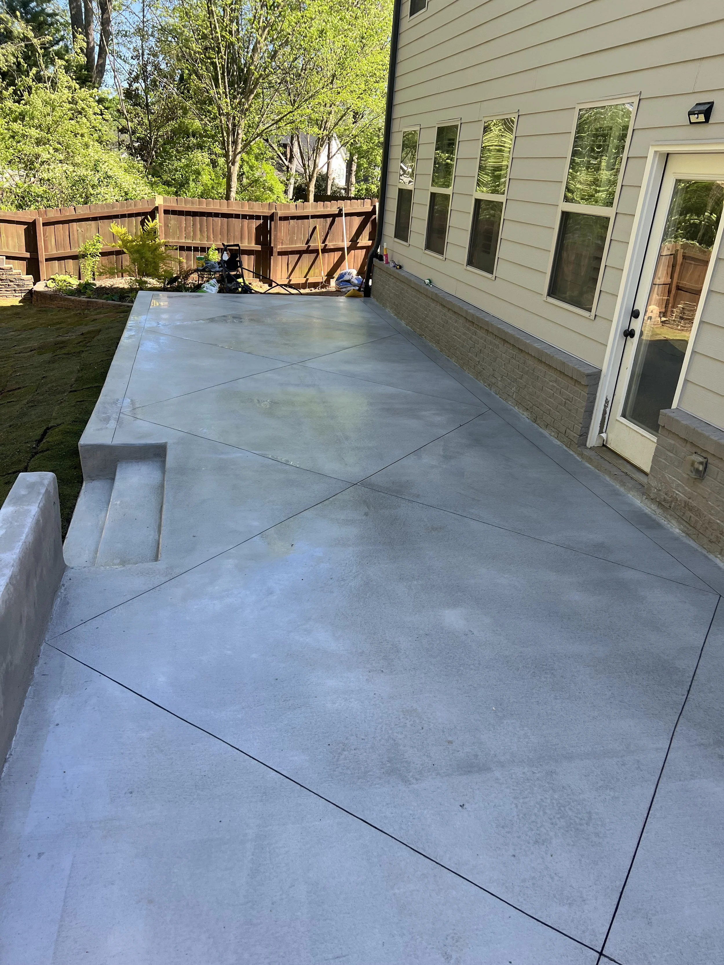Finished concrete patio with steps near house with siding, windows, and a door, fenced backyard with trees and plants, and outdoor tools in the background.