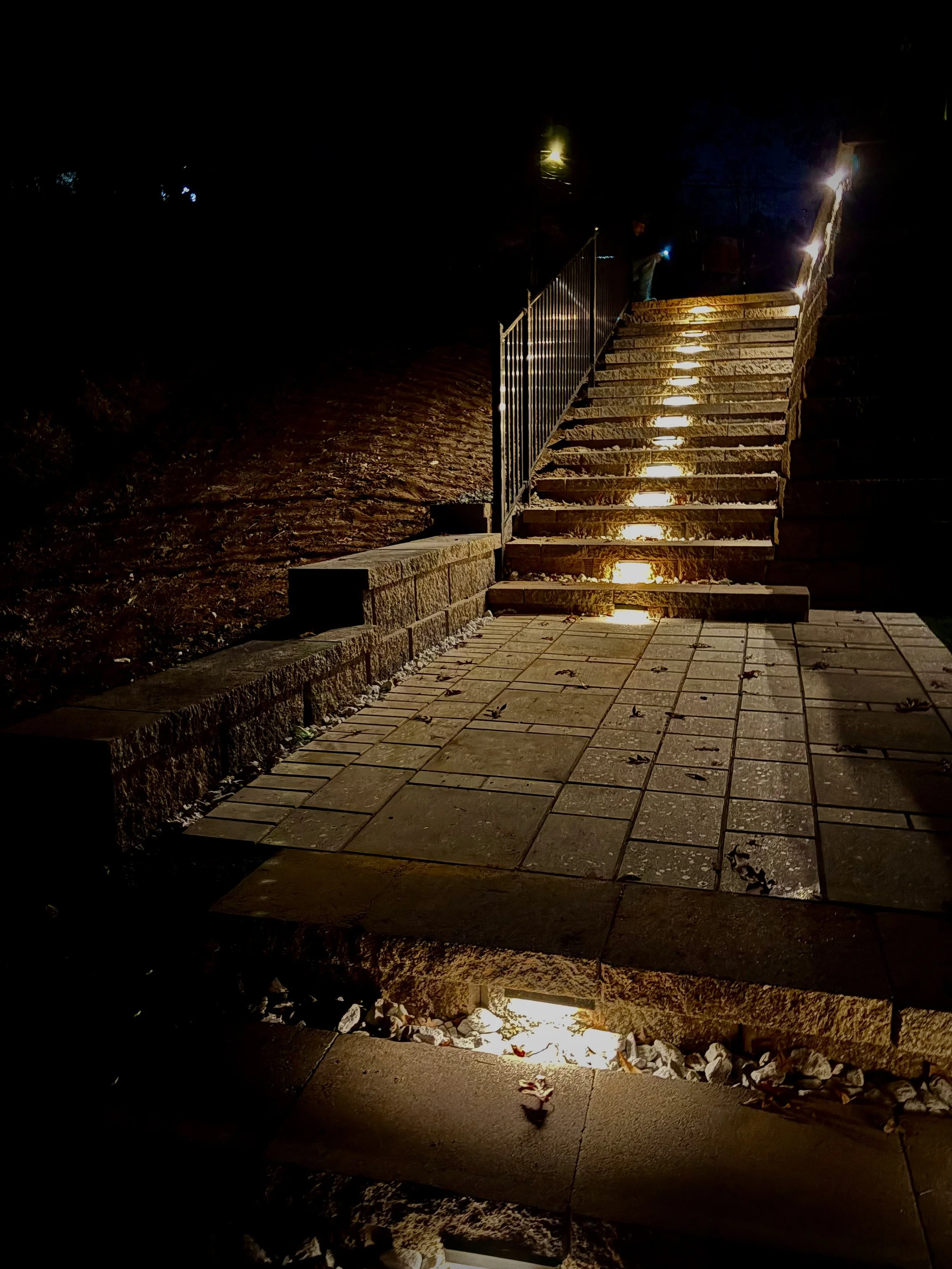 Outdoor staircase illuminated by pathway lights at night, with metal railing on one side and stone pavers at the base.
